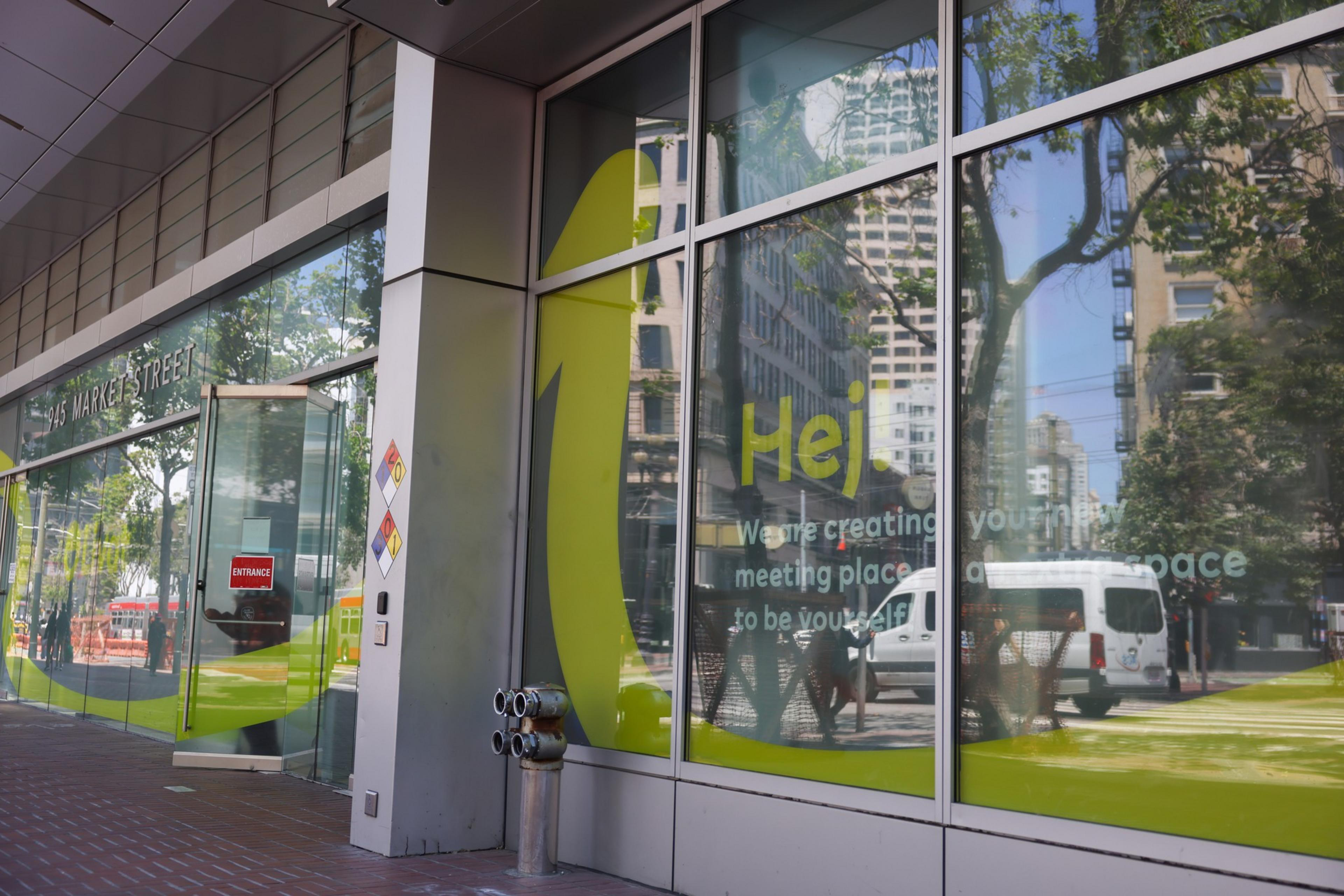 A glass building facade reflects city buildings and trees, with yellow text on the window reading "Hej" and a message about creating a meeting space.