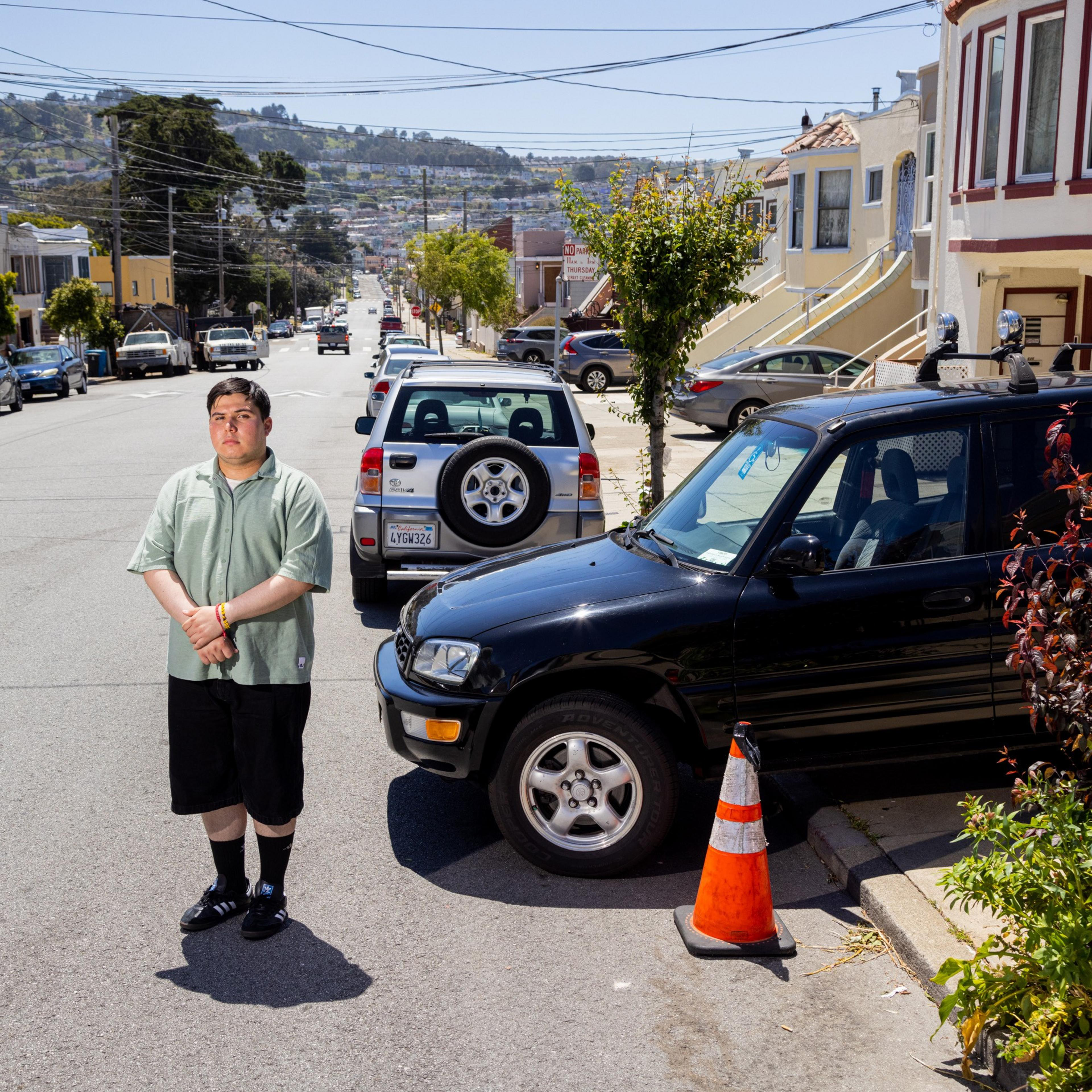 A man stands on a sunny street with parked cars, colorful houses, hills in the background, and a traffic cone nearby.