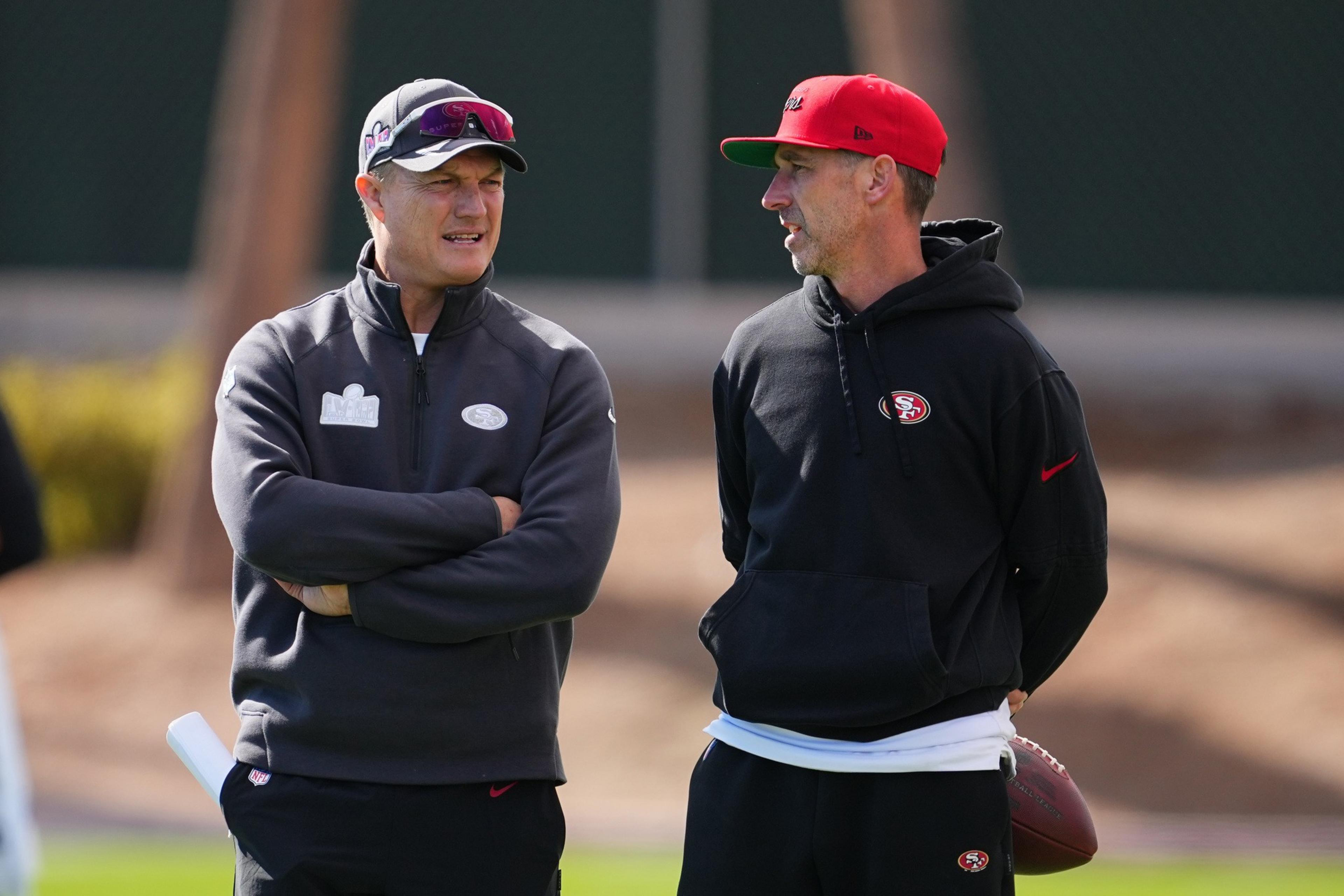 Two men are standing outside, wearing sports attire with the San Francisco 49ers logo. One has arms crossed, the other holds a football, both in conversation.