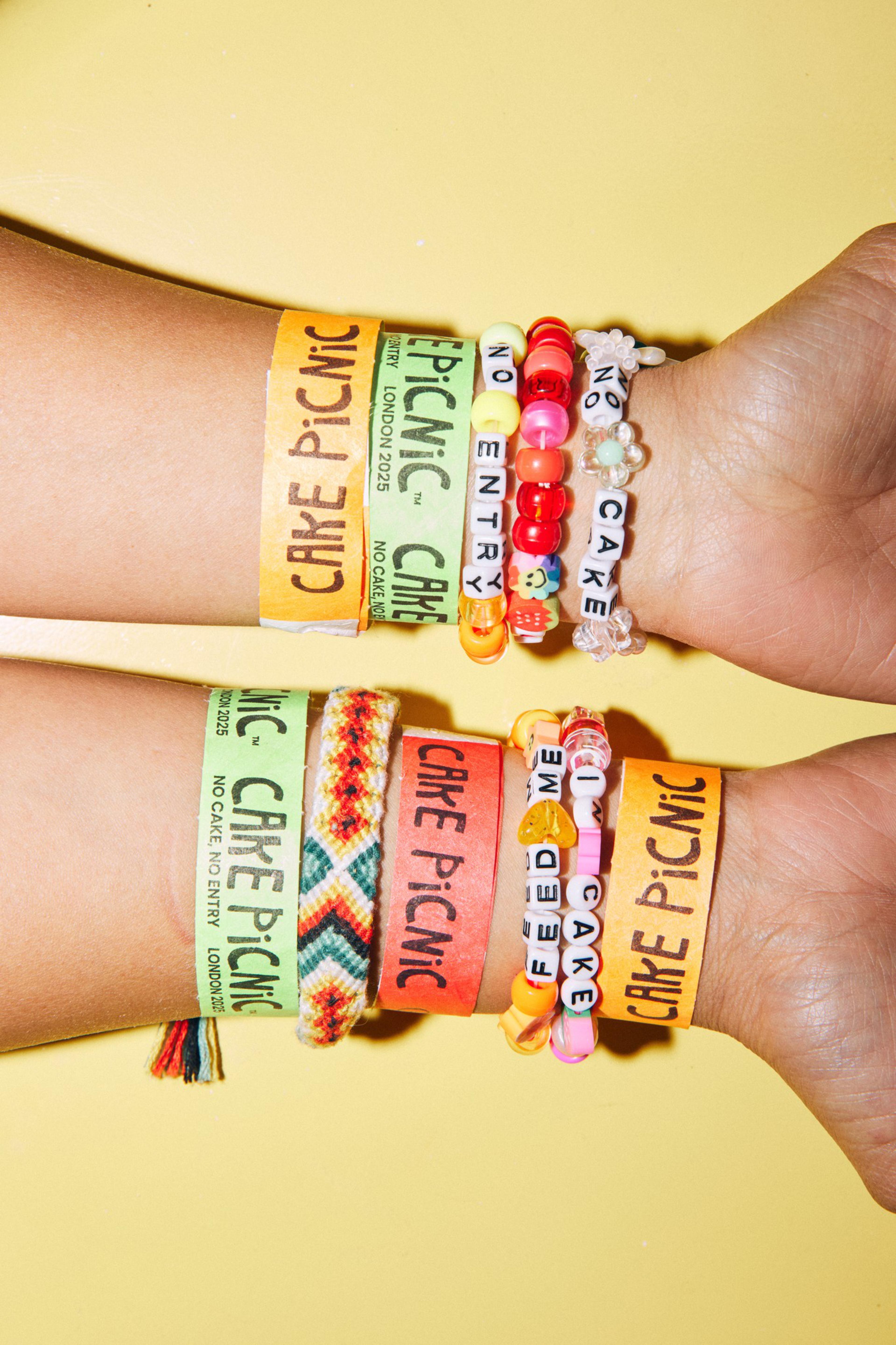 Two wrists with various colorful bracelets and paper wristbands that say “CAKE PICNIC” and “NO CAKE NO ENTRY” on a yellow background.