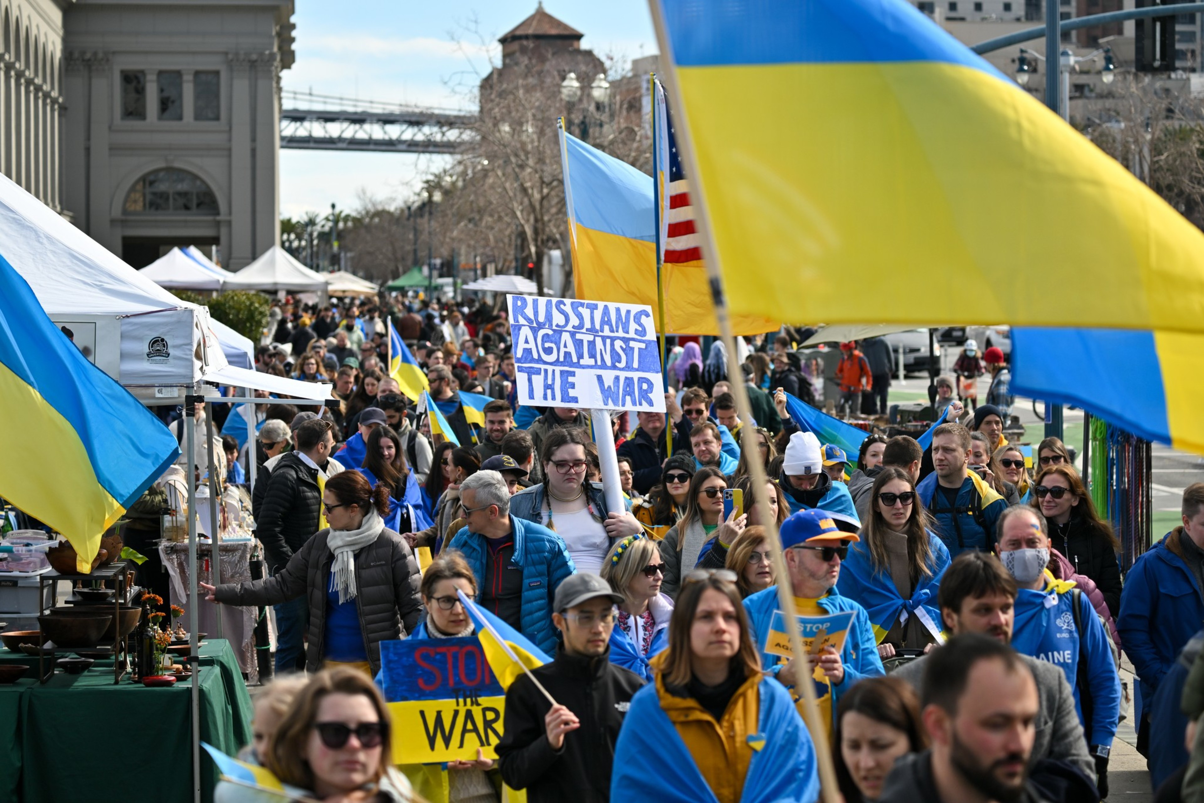 A diverse group of people march in a crowded street, holding Ukrainian flags and anti-war signs, with tents and a bridge visible in the background.