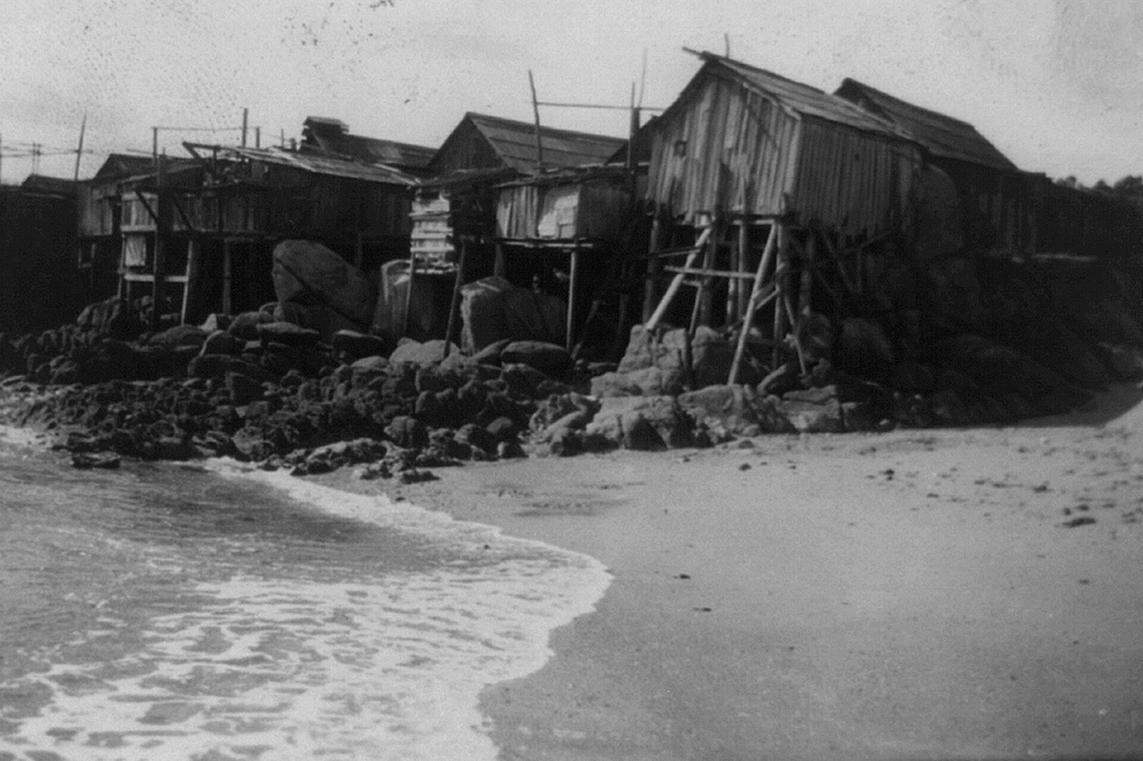 A row of weathered wooden shacks built on rocky stilts lines a sandy beach with gentle waves approaching the shore.