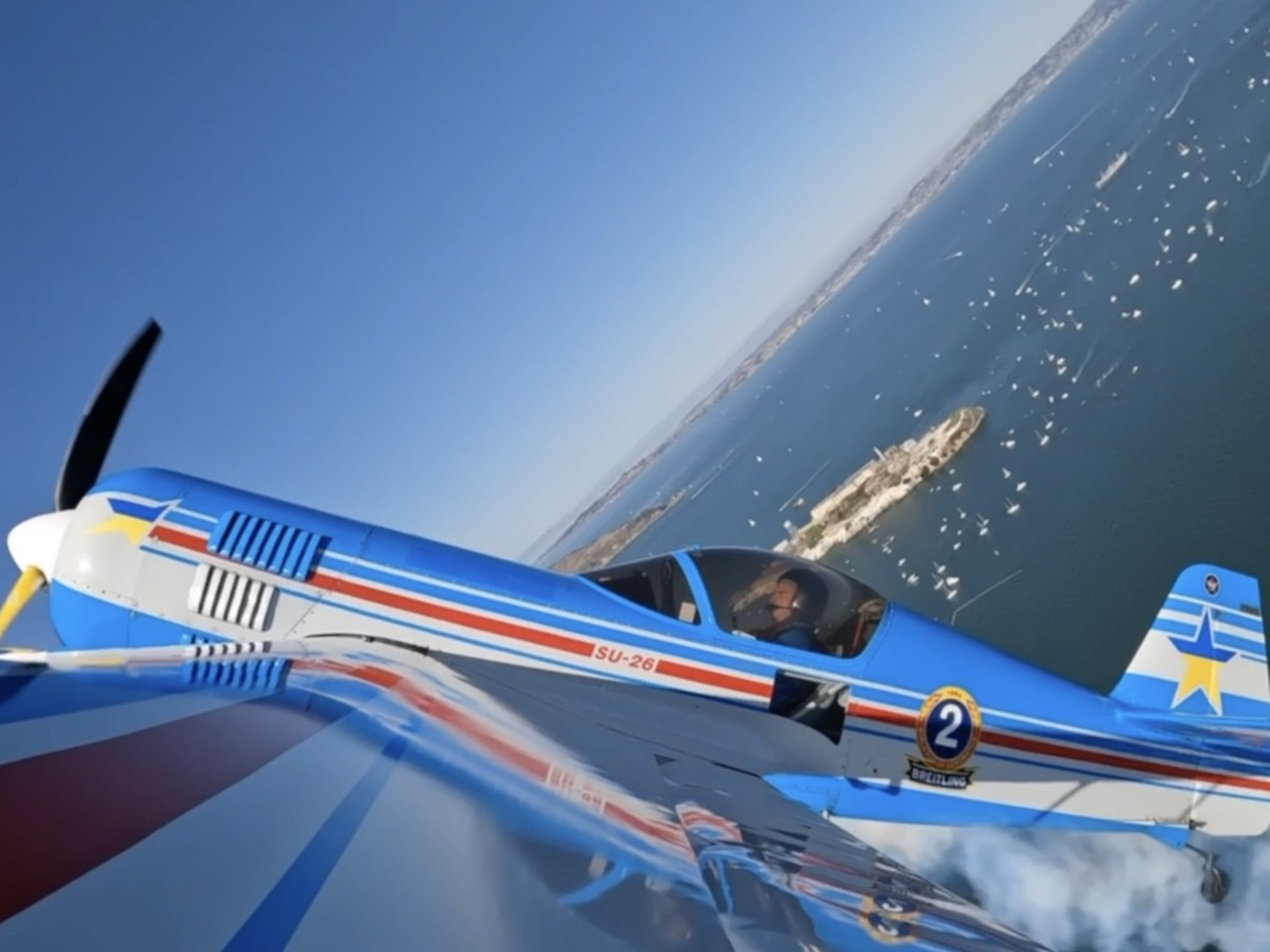 A blue SU-26 stunt plane with red, white, and blue stripes flies over water filled with many boats near a small island, with the pilot visible inside.