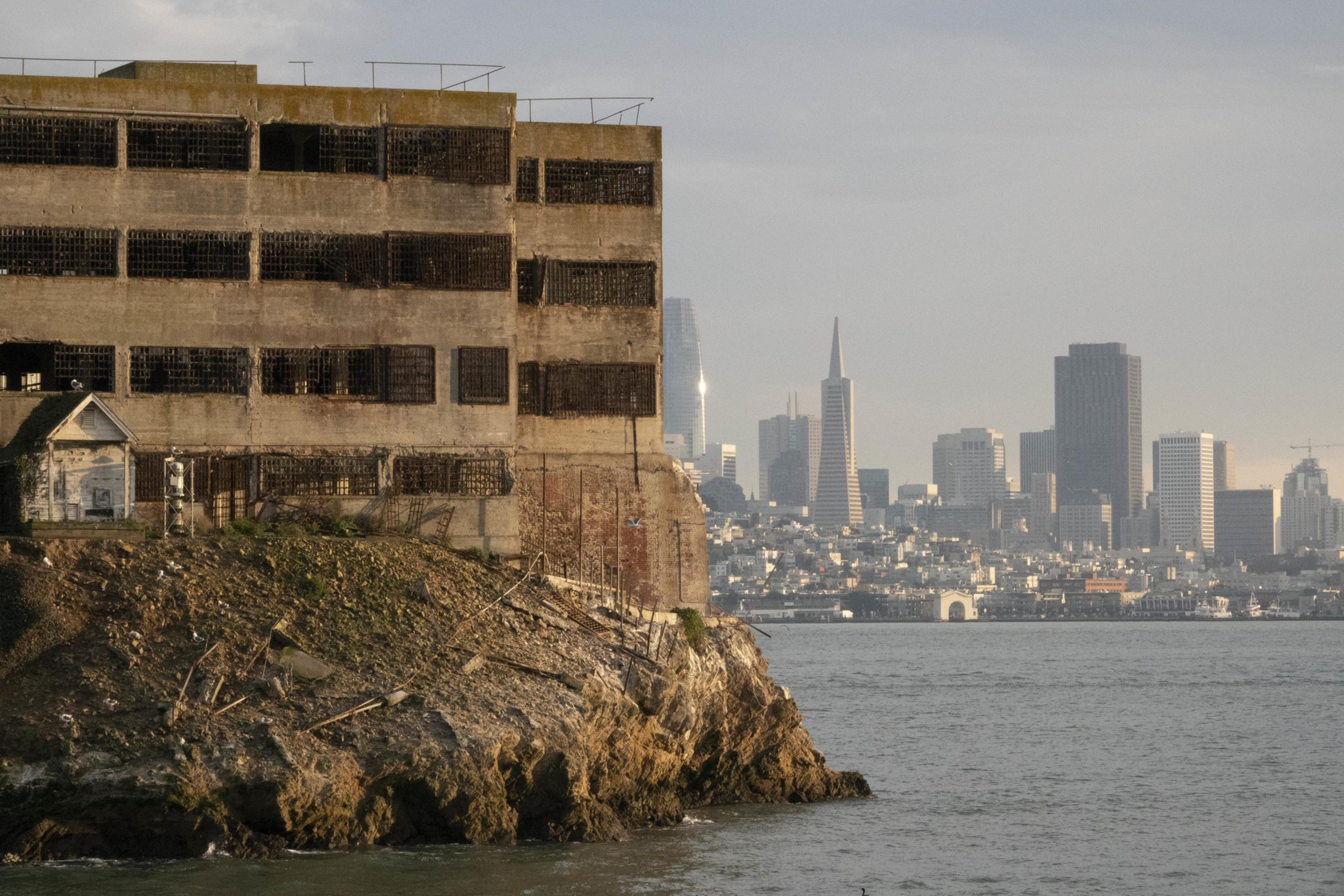 A weathered industrial building with barred windows stands by the water. In the background, a city skyline with high-rise buildings is visible.