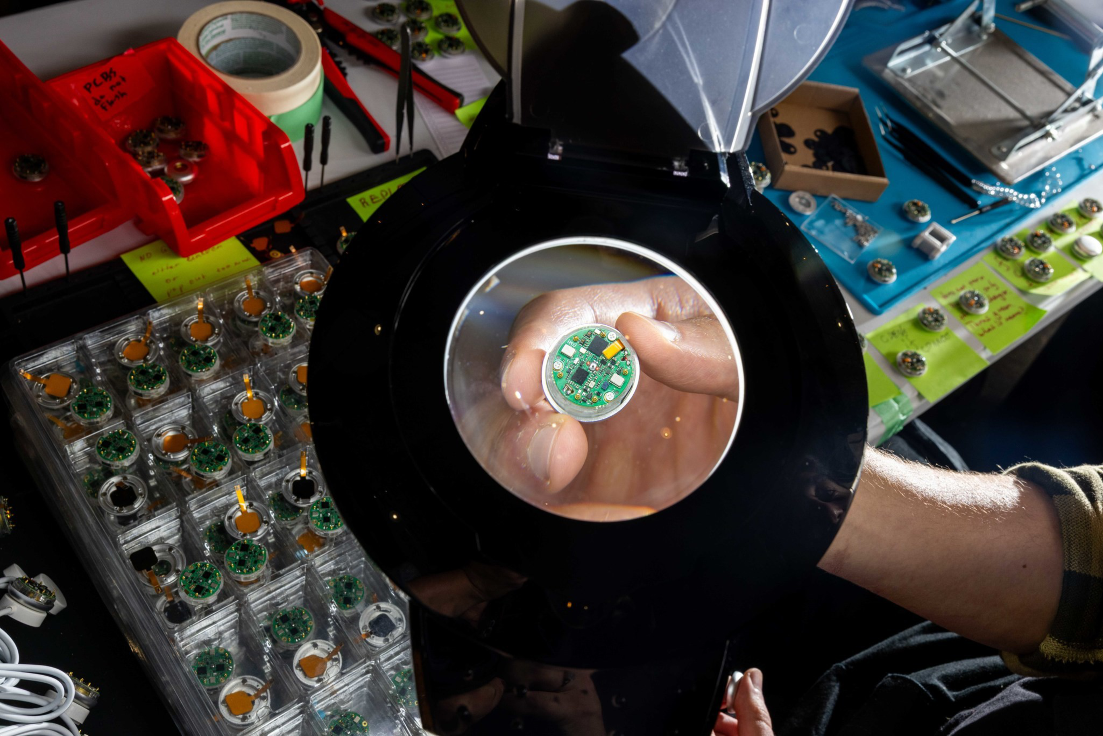 A hand holds a small green circuit board under a magnifying glass, with multiple similar circuit boards and tools spread on a table.