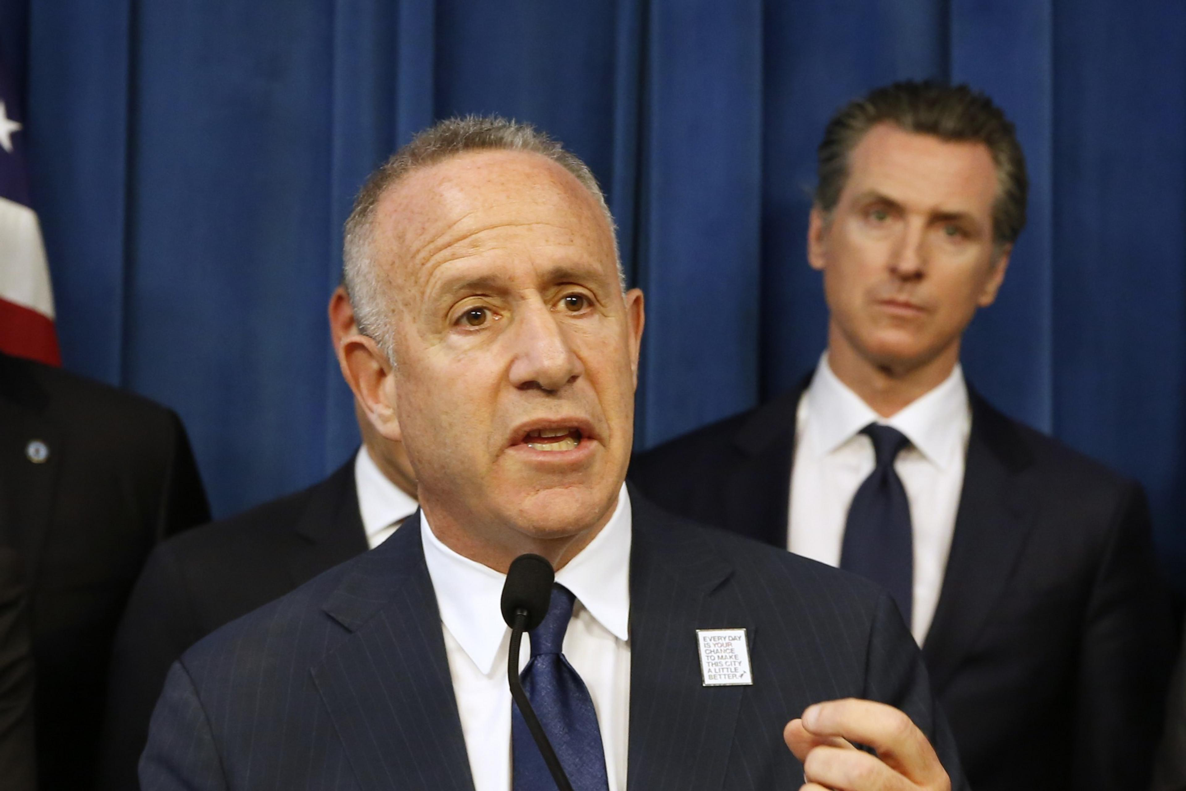 Man wearing blazer and blue tie speaks into microphone as another man looks on behind him