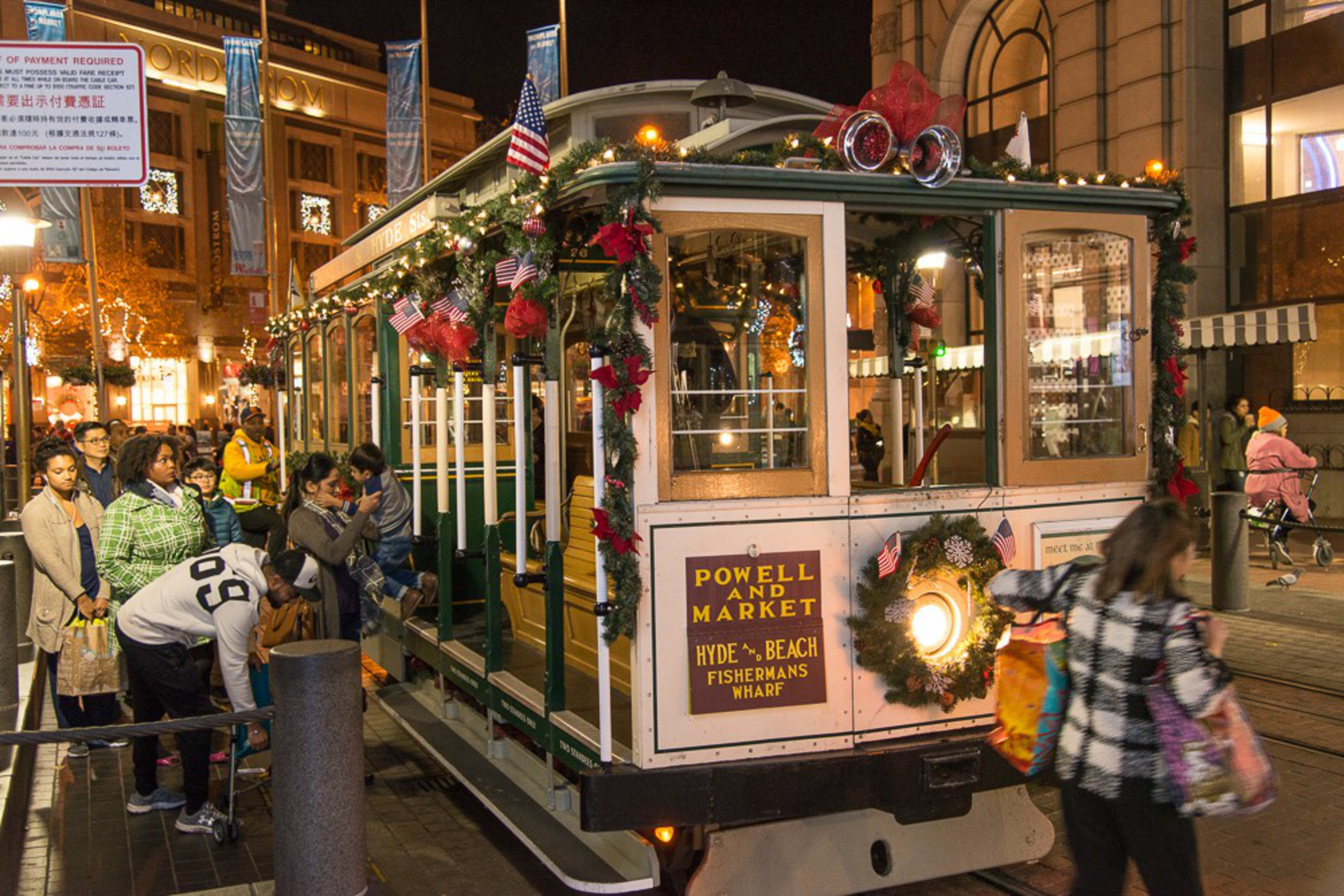 A decorated San Francisco cable car with holiday wreaths and garlands waits at a stop while people stand in line, with city lights glowing in the background.