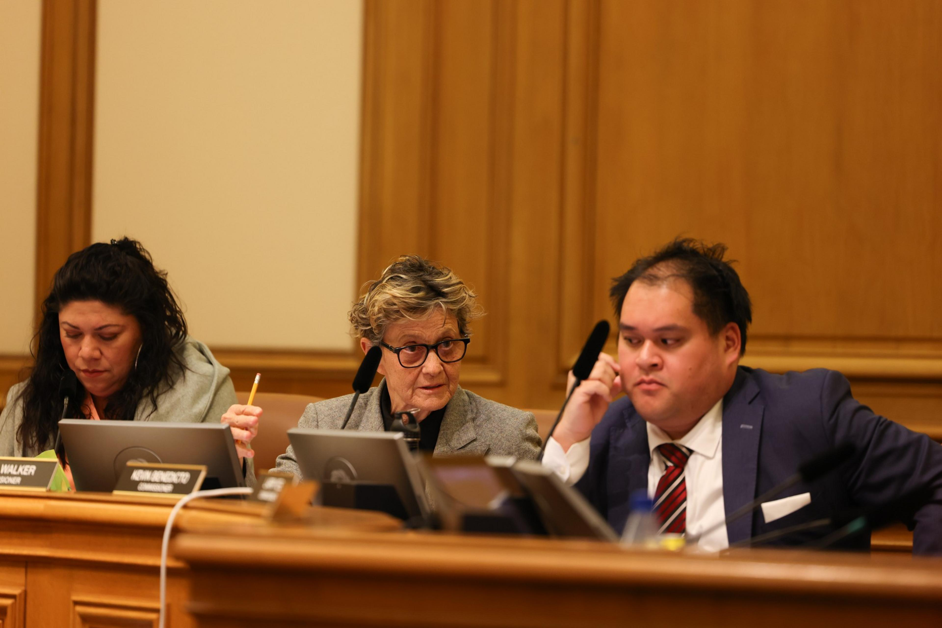 Three people sit at a wooden panel desk with microphones and nameplates, engaged in discussion during a formal meeting or hearing.