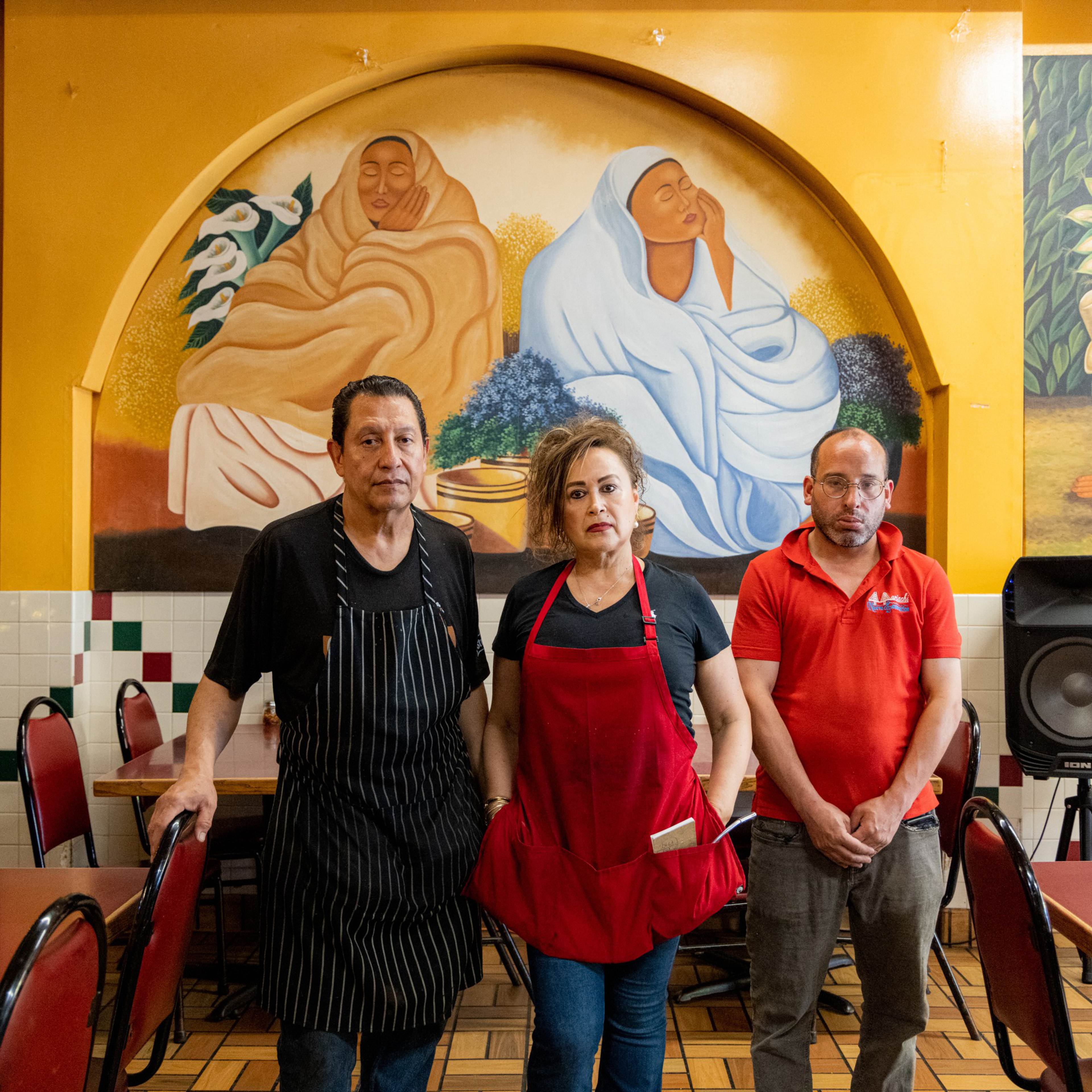 Three people stand in front of a mural inside a vividly decorated restaurant. Two wear aprons and the third, in a red shirt, stands beside them.