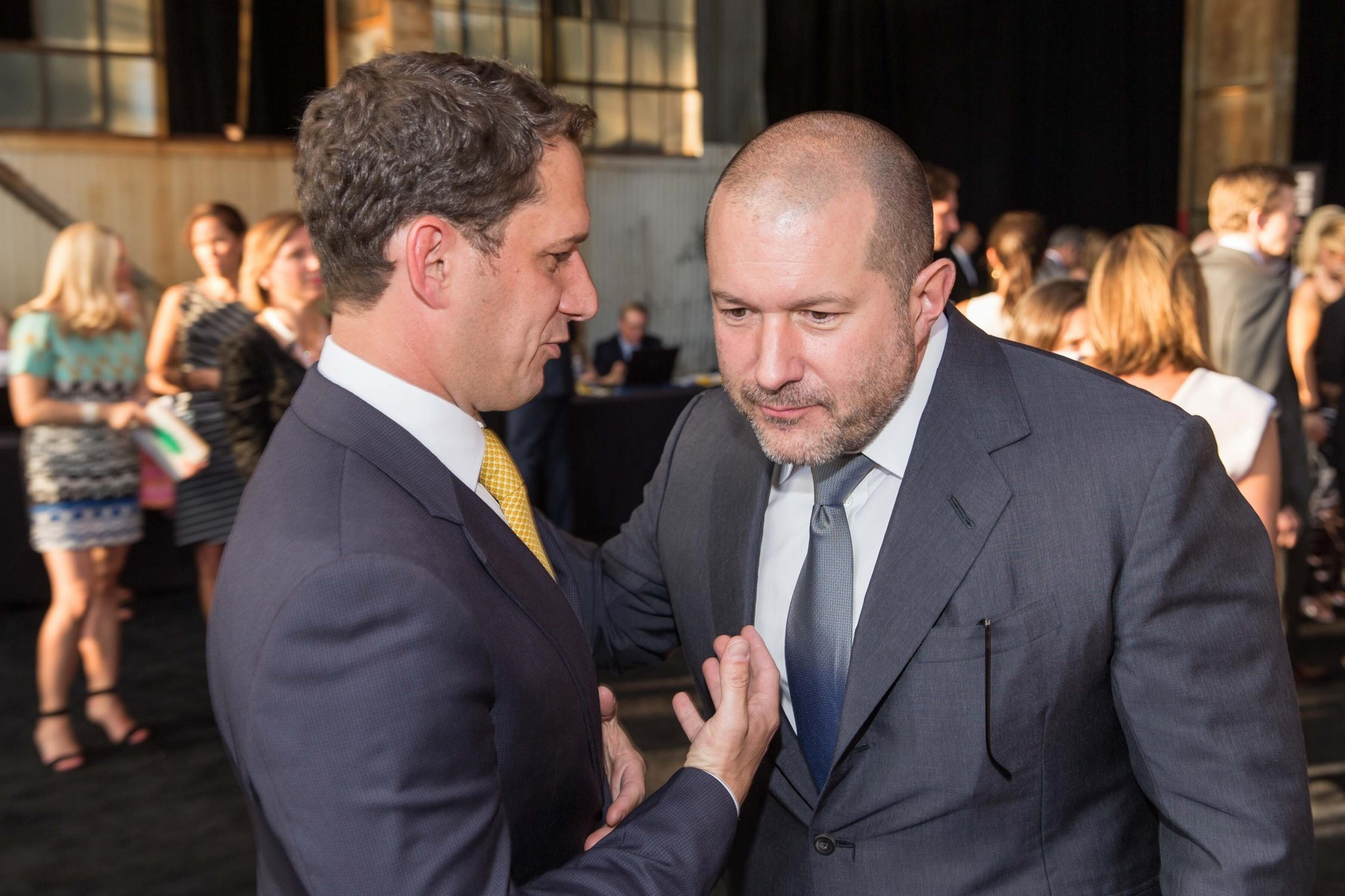Two men in suits are engaged in conversation at a social event, surrounded by people in formal attire. The lighting is warm and the setting appears lively.