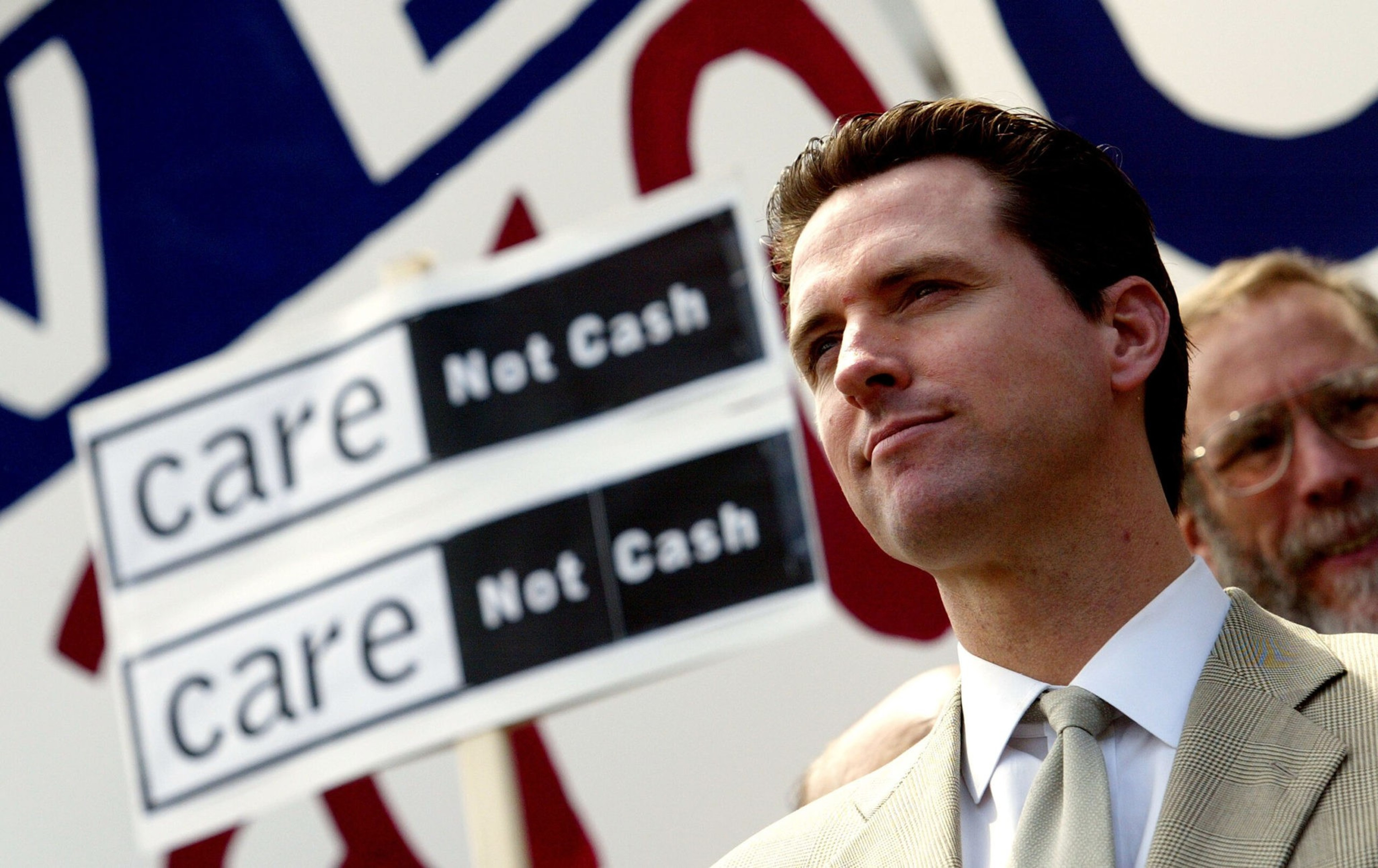 A man in a light suit and tie stands confidently near two protest signs reading "care Not Cash" against a blurred background.