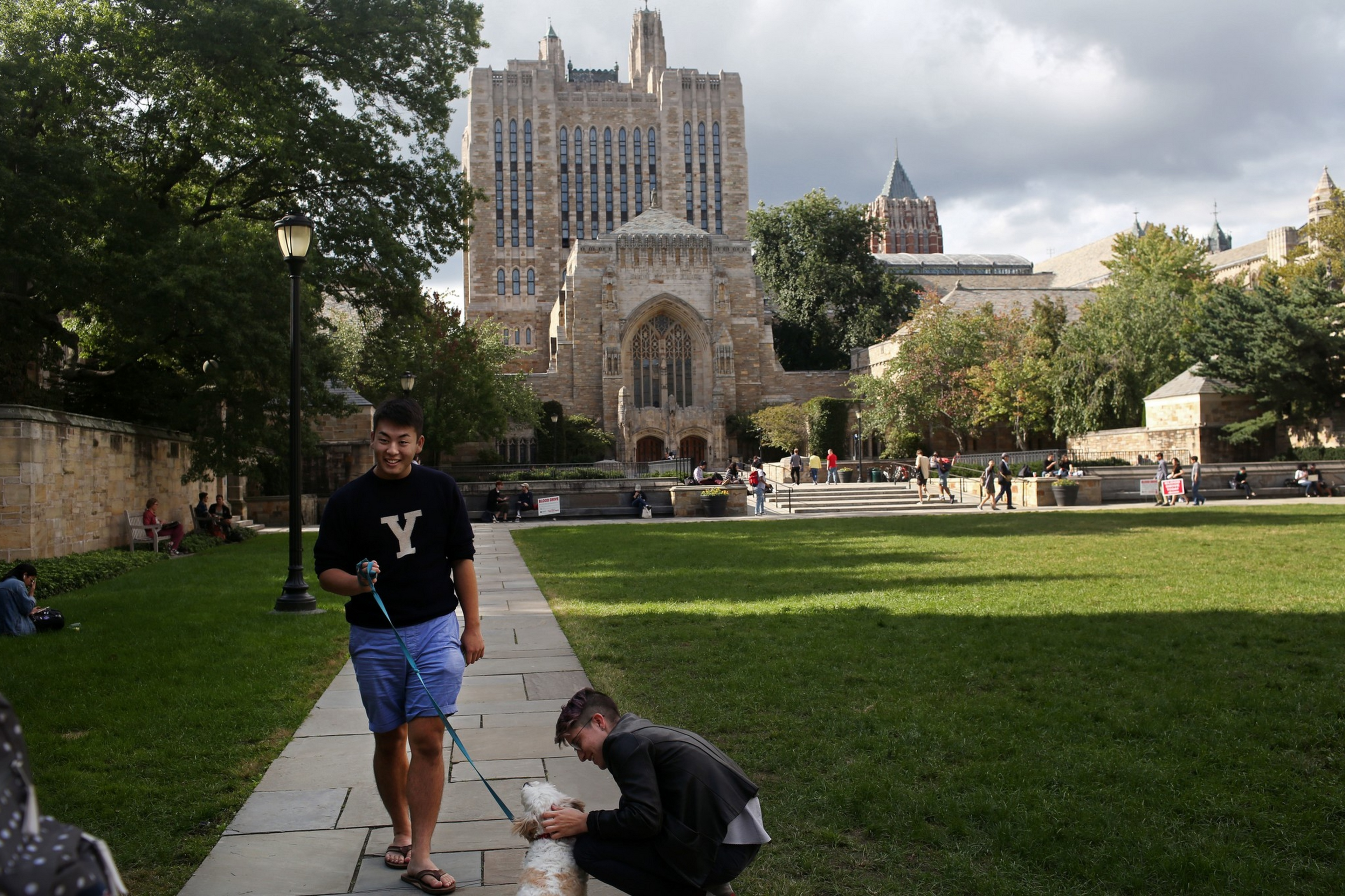 A man in a black "Y" sweatshirt walks a white dog on a leash while another person crouches to pet the dog on a path in front of a large Gothic building.