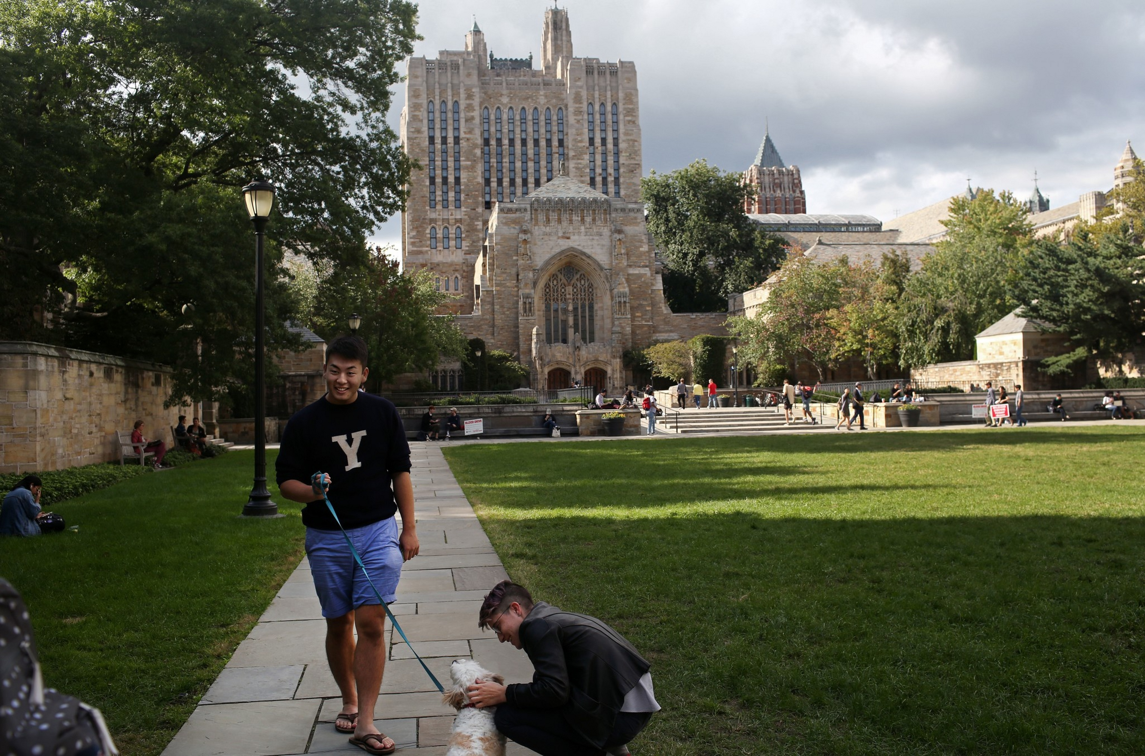 A man in a black "Y" sweatshirt walks a white dog on a leash while another person crouches to pet the dog on a path in front of a large Gothic building.
