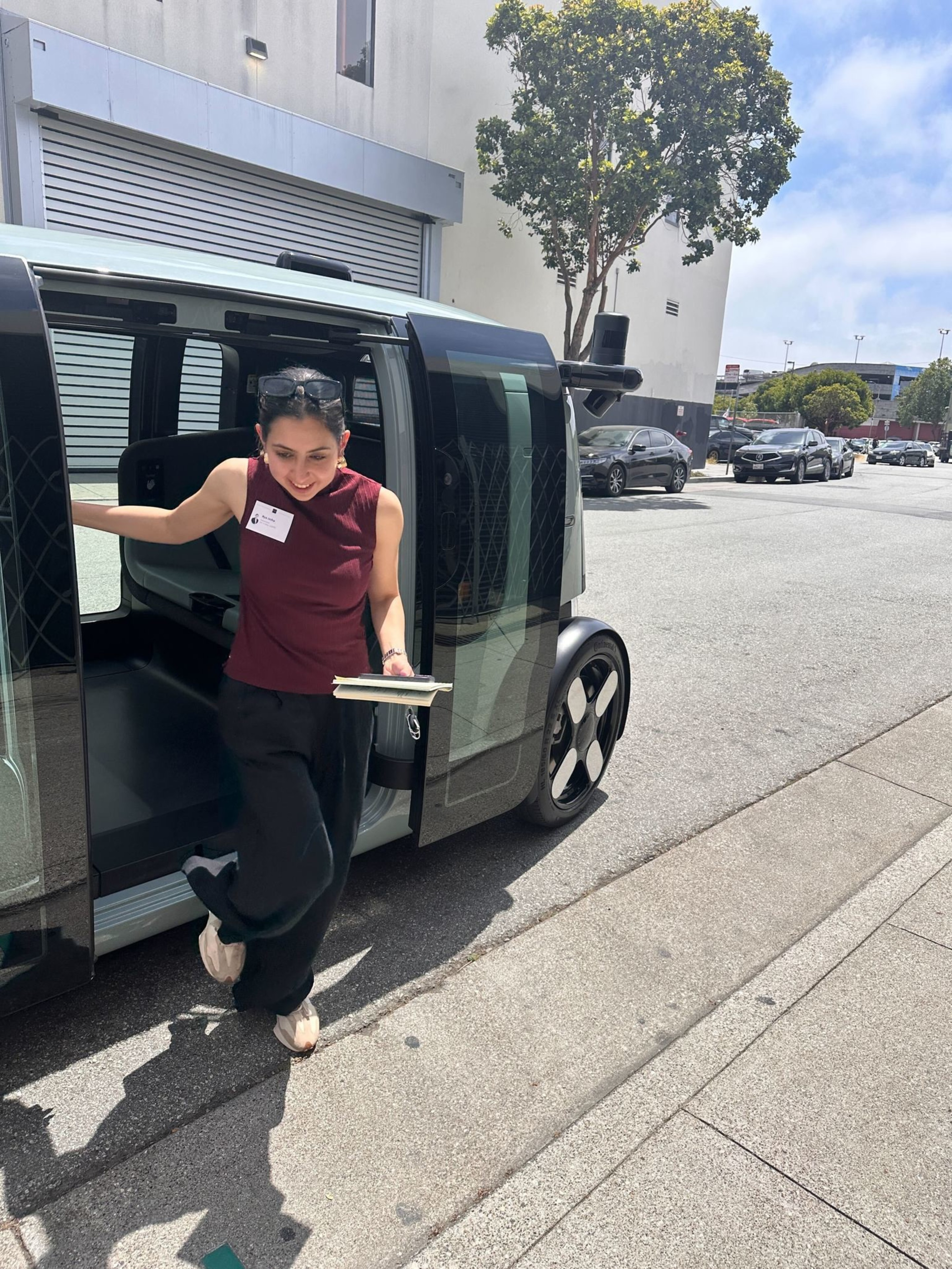 A woman steps out of a modern, boxy vehicle on a sunny street. She holds a notebook and wears a name tag. Trees and parked cars line the road.
