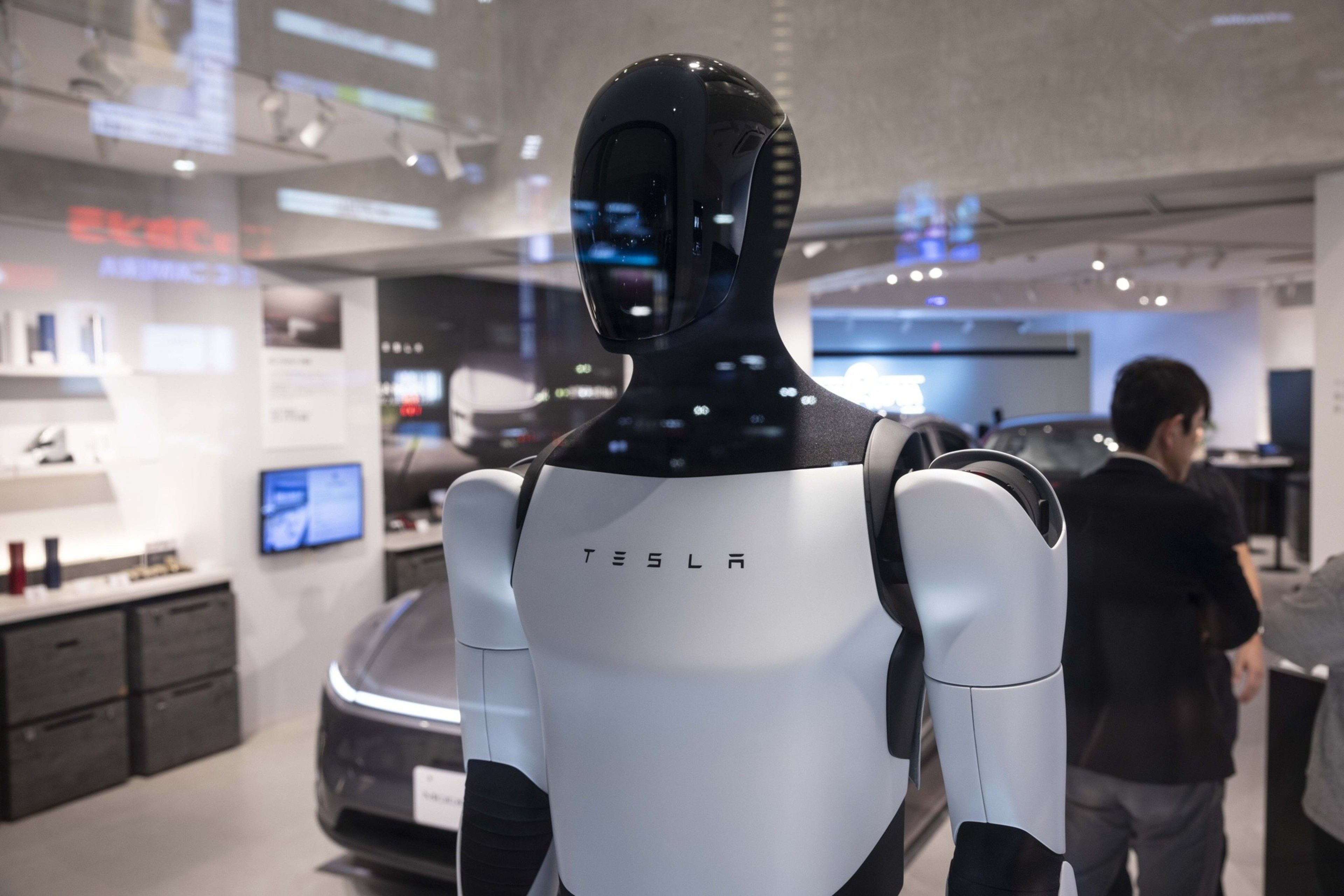 A black and white humanoid robot with "TESLA" on its chest stands indoors, facing slightly to the right, with people and a car in the background.