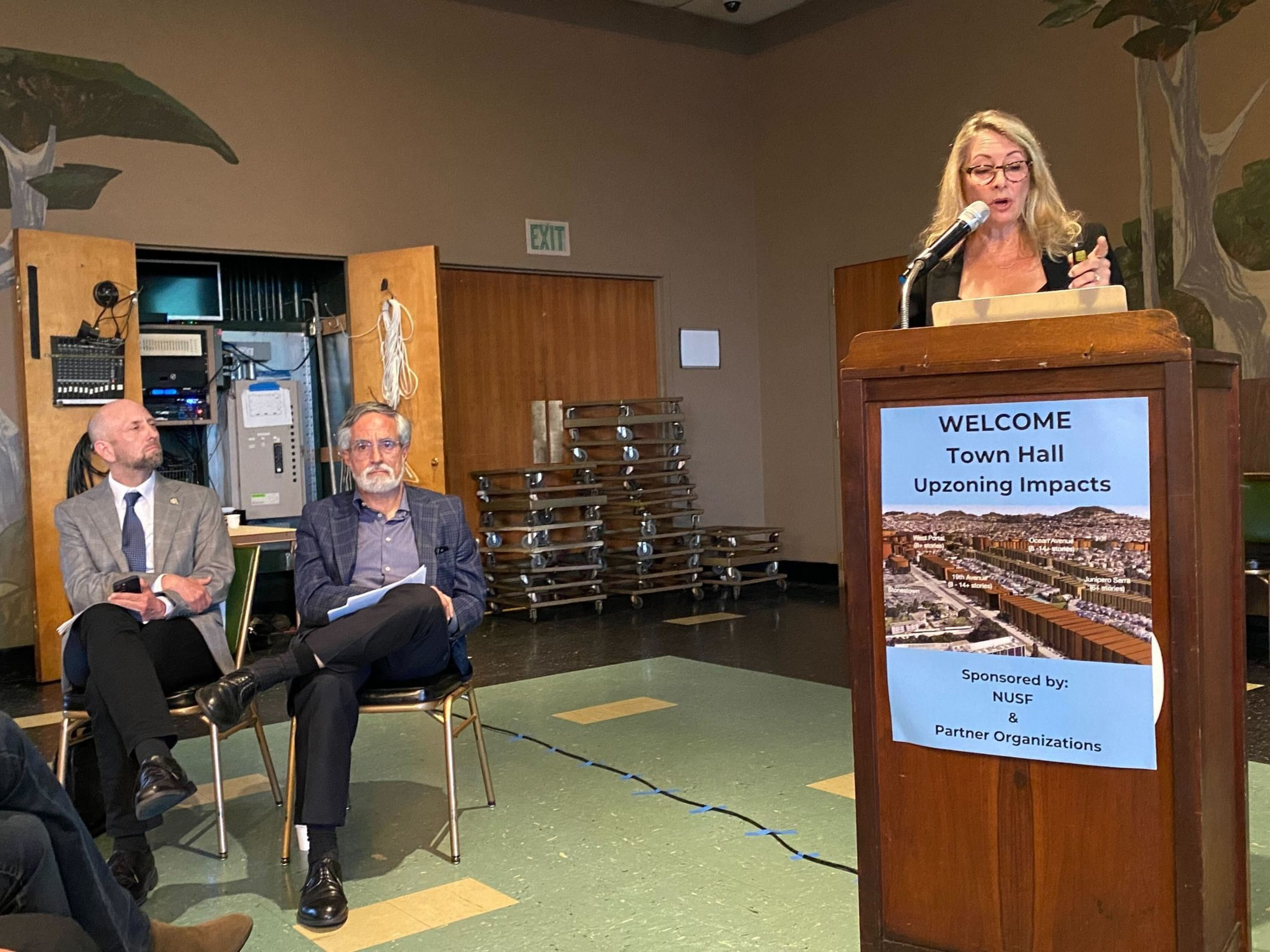 A woman speaks at a podium labeled &quot;Town Hall Upzoning Impacts.&quot; Two men sit beside her, attentively listening in a room with a wooden floor and technical equipment in the background.