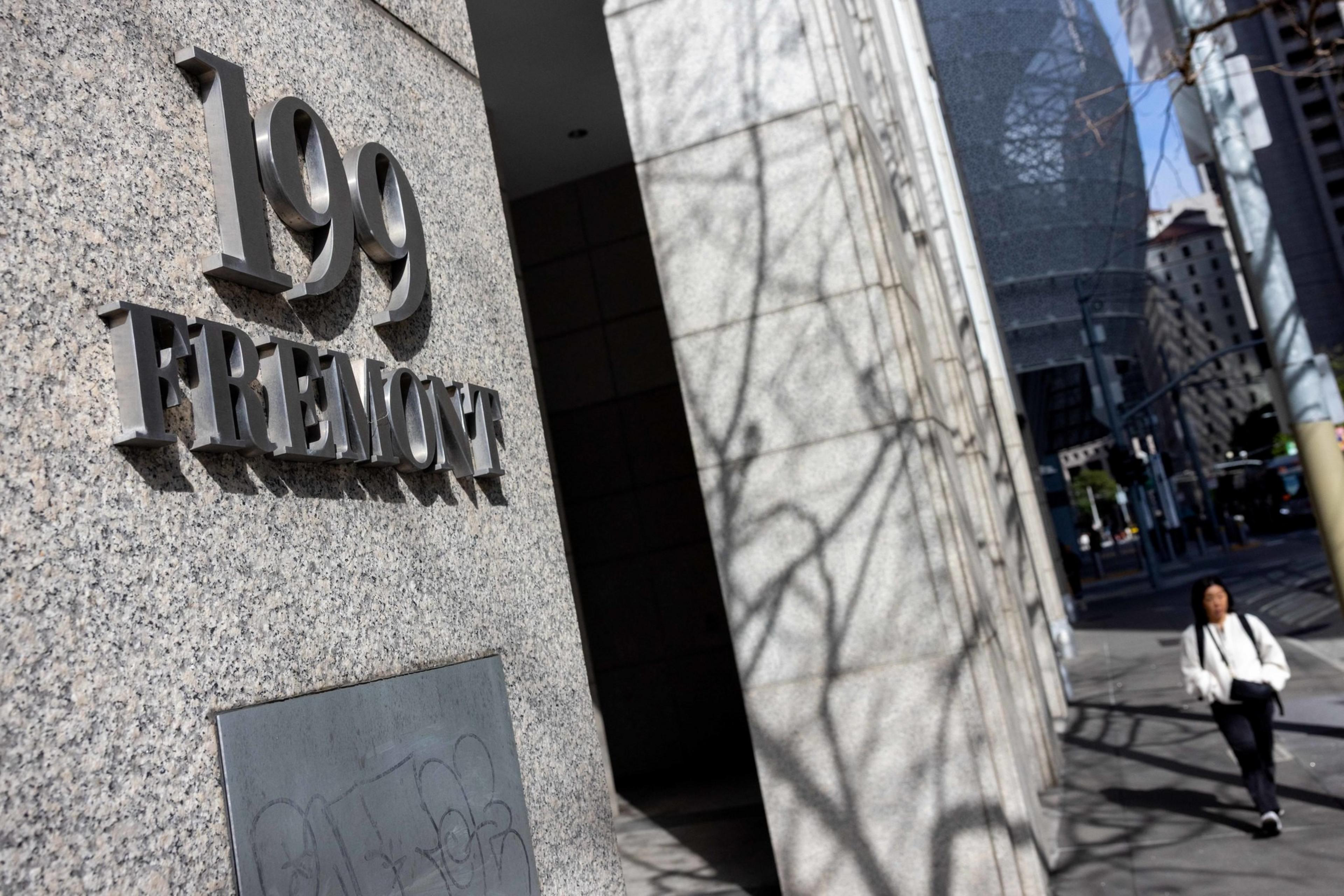 The image shows a building entrance with "199 Fremont" displayed in metal letters on a stone wall, while a person walks on the sidewalk nearby. Shadows of branches are visible on the wall.
