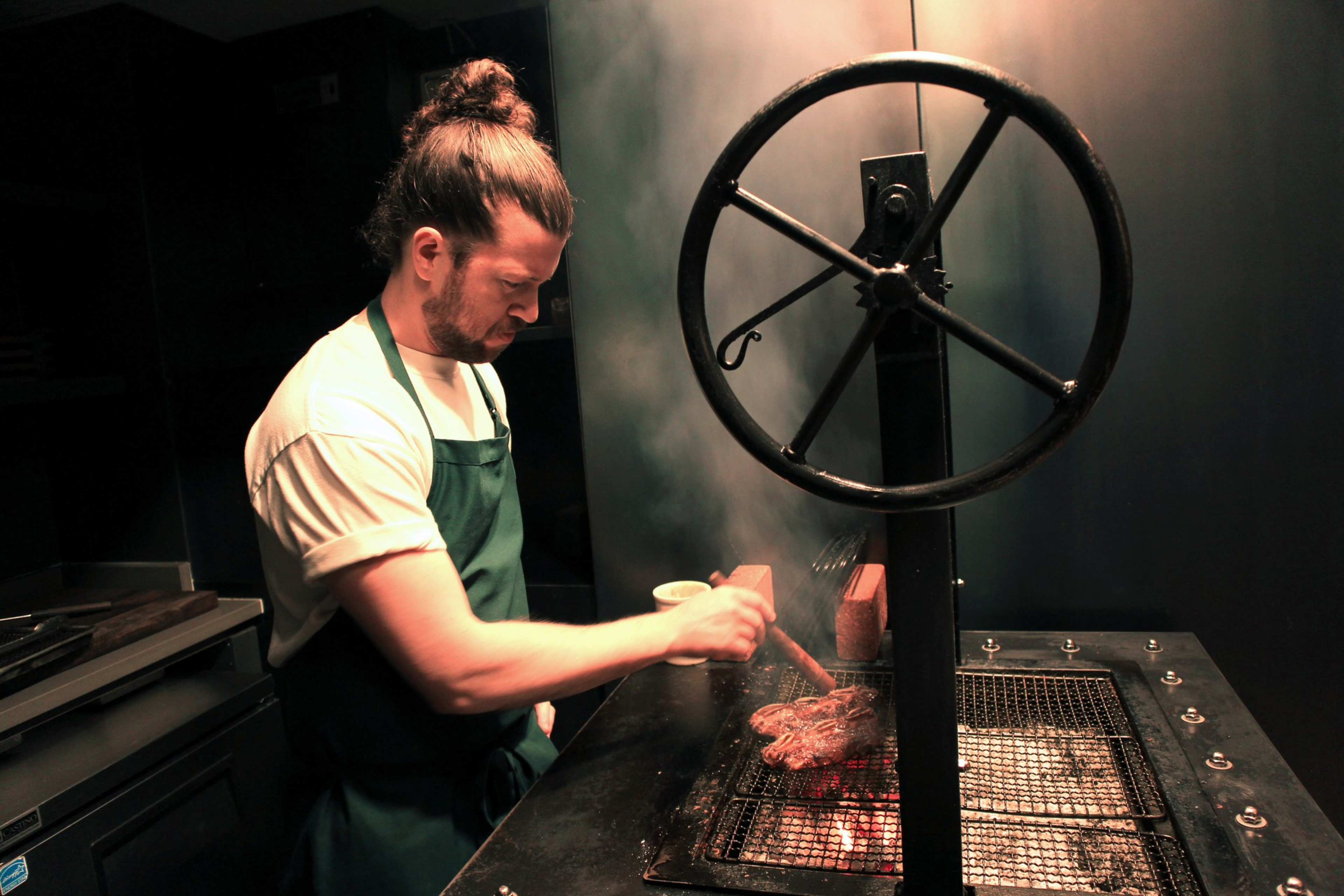 A person with a bun and apron grills meat over an open flame, using tongs. Smoke rises as they focus on cooking in a dimly lit space.