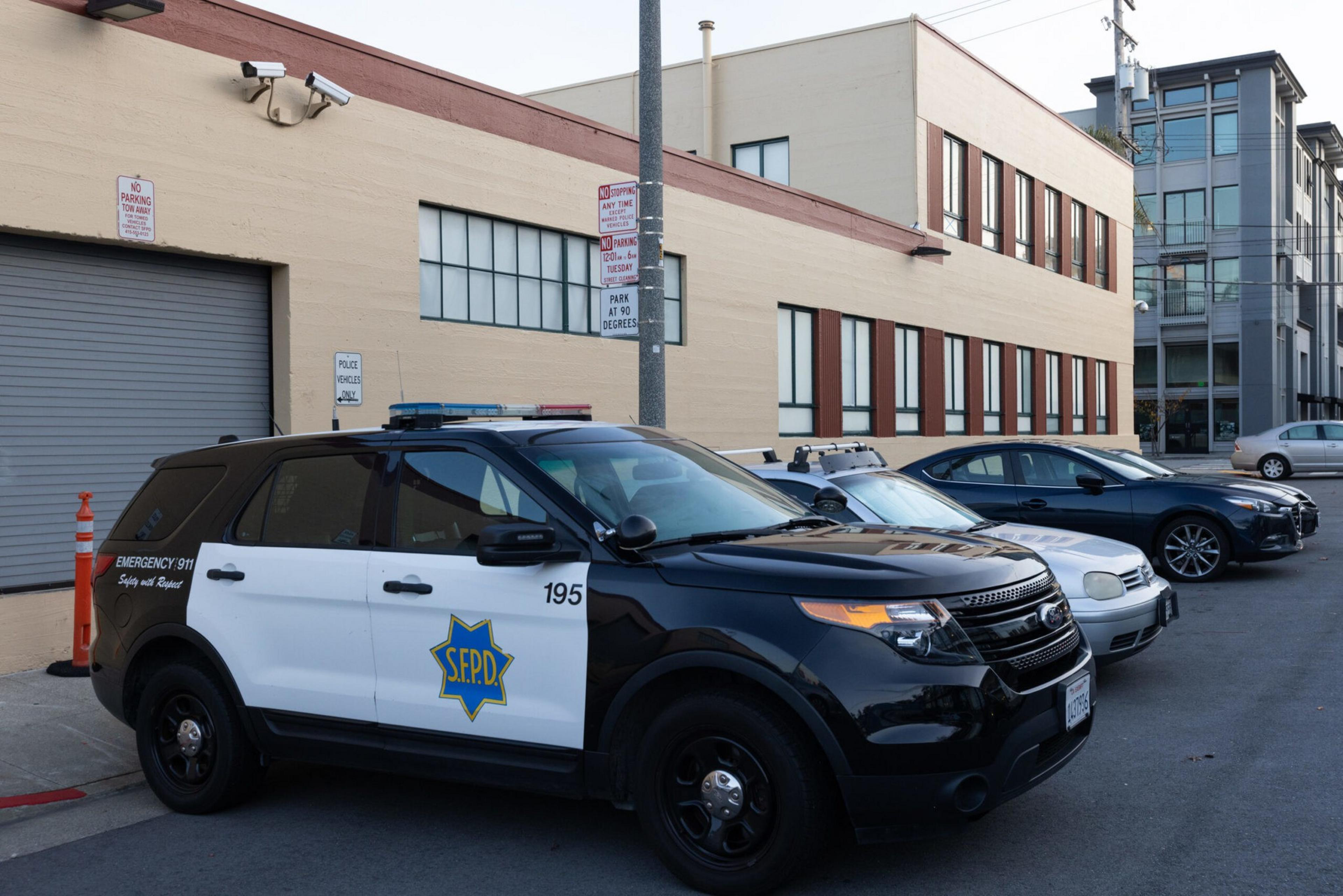 A black and white San Francisco police SUV is parked next to a white car and a dark blue sedan along a beige building with large windows.