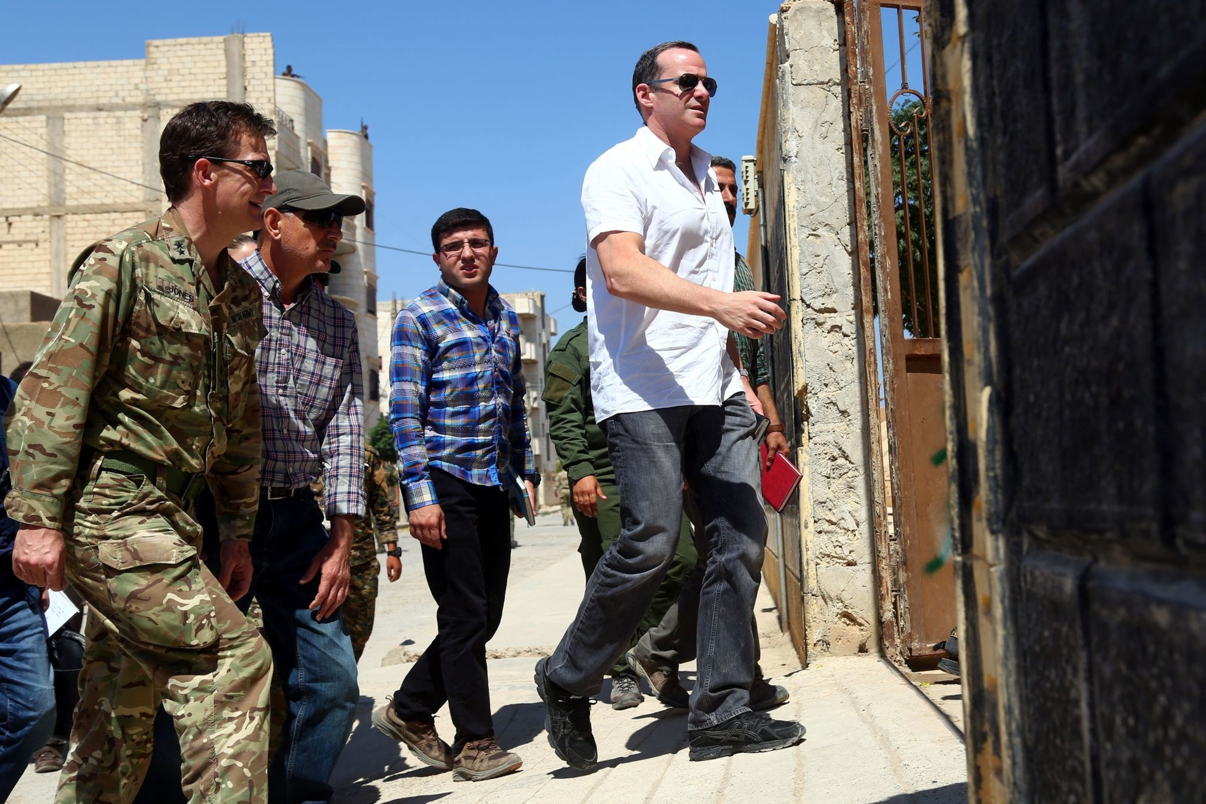 A group of men, some in military clothing and others in casual wear, walk together on a sunlit street with buildings and a gate.