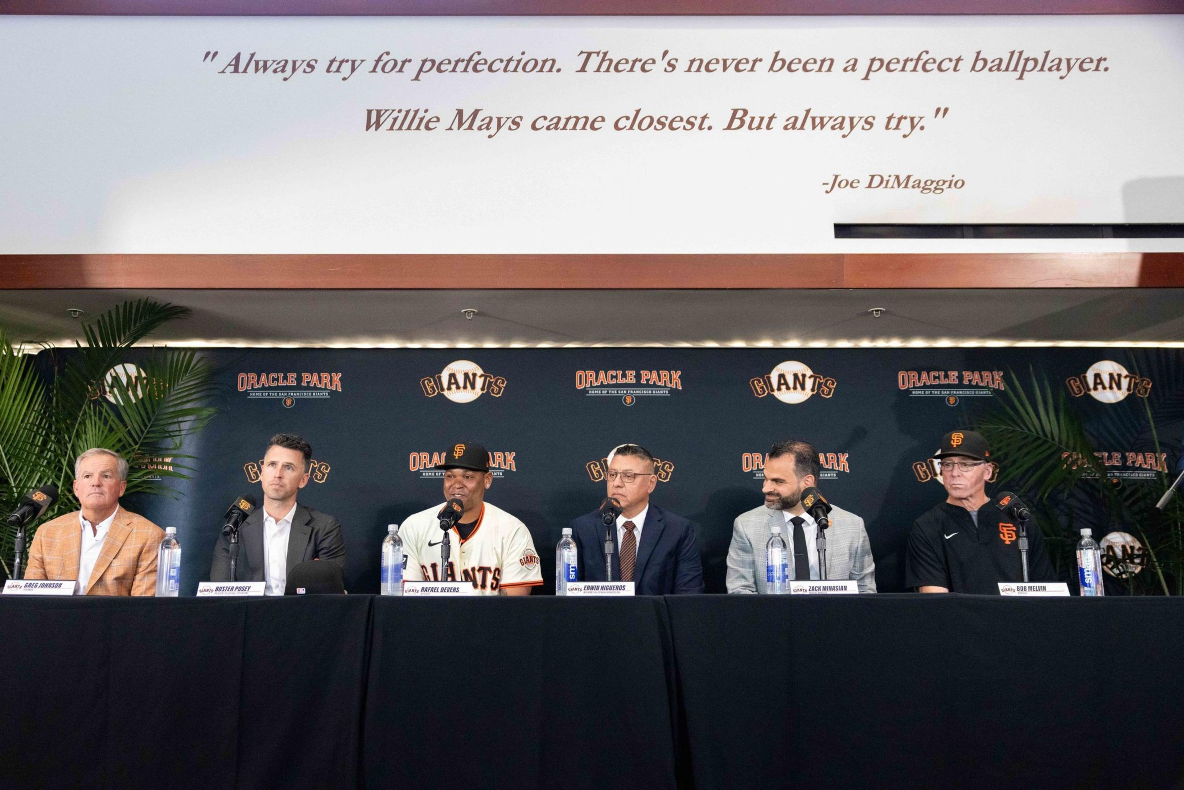 Six men sit at a table with microphones in front of a Giants backdrop, under a quote by Joe DiMaggio. Some are in suits, and one wears a Giants jersey.