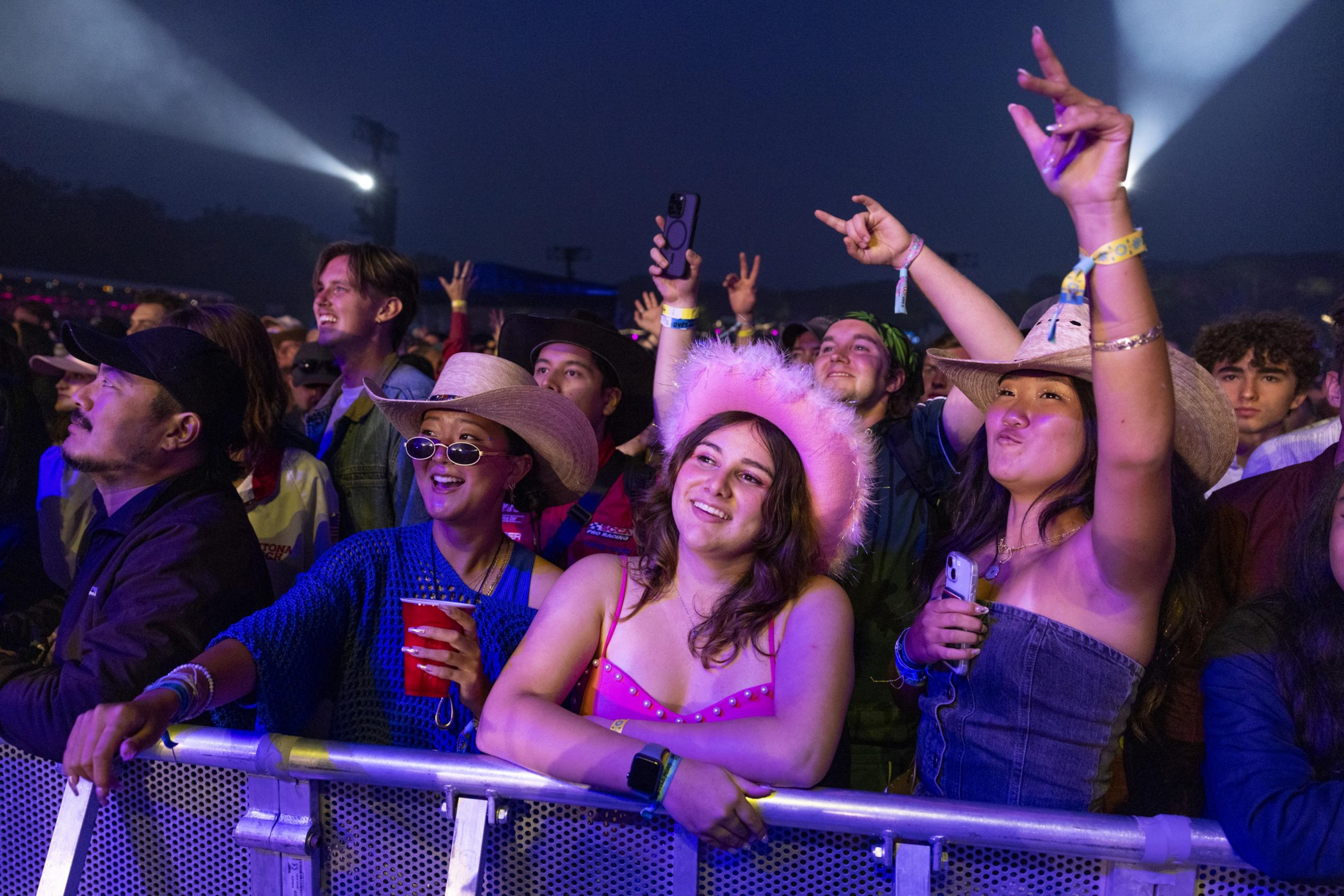 A lively crowd at an outdoor concert enjoys the music. People in colorful outfits and hats are cheering, smiling, and raising their hands under bright stage lights.