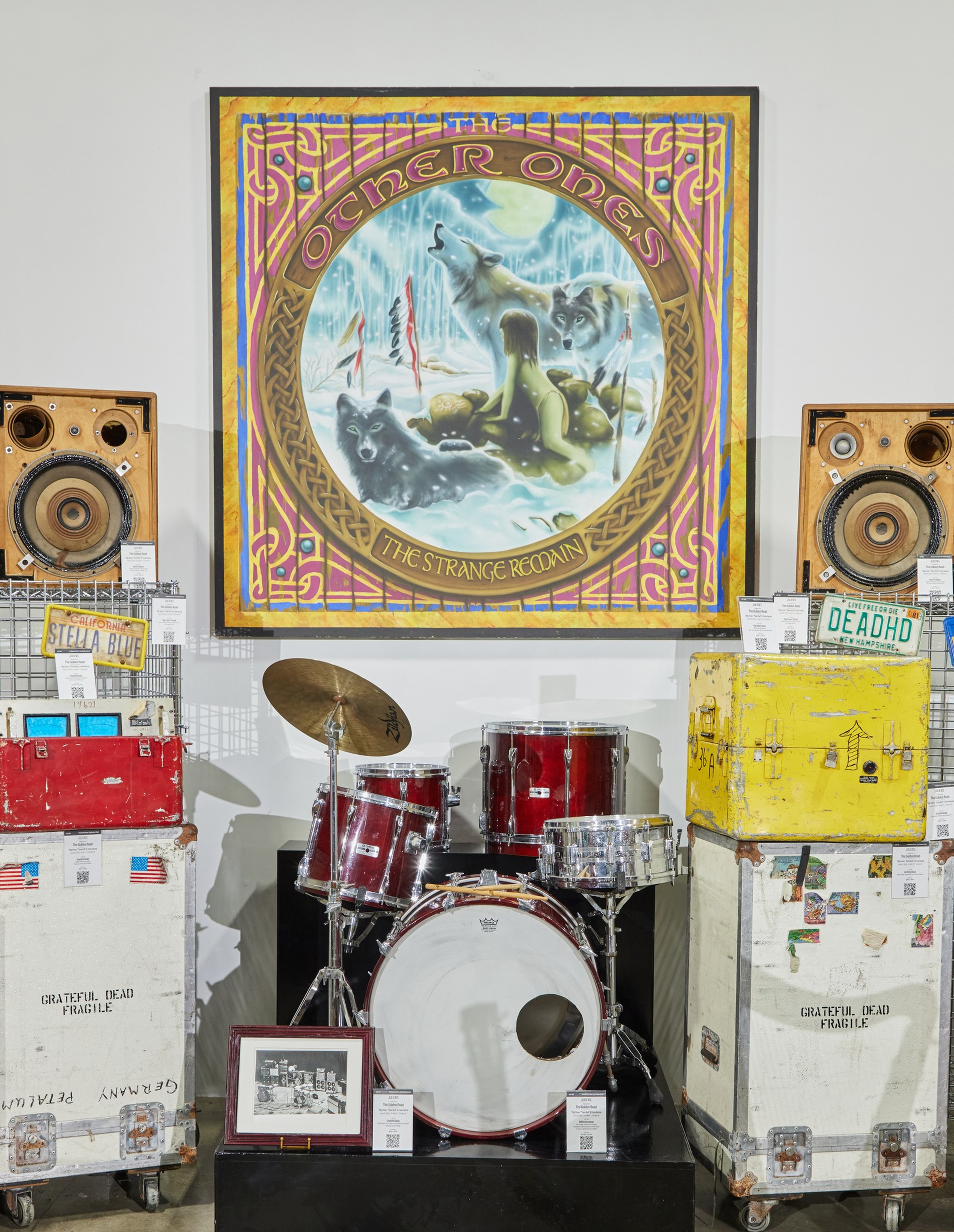 A framed artwork of wolves howling in snow hangs above a red drum set surrounded by vintage speakers and travel cases labeled “Grateful Dead.”