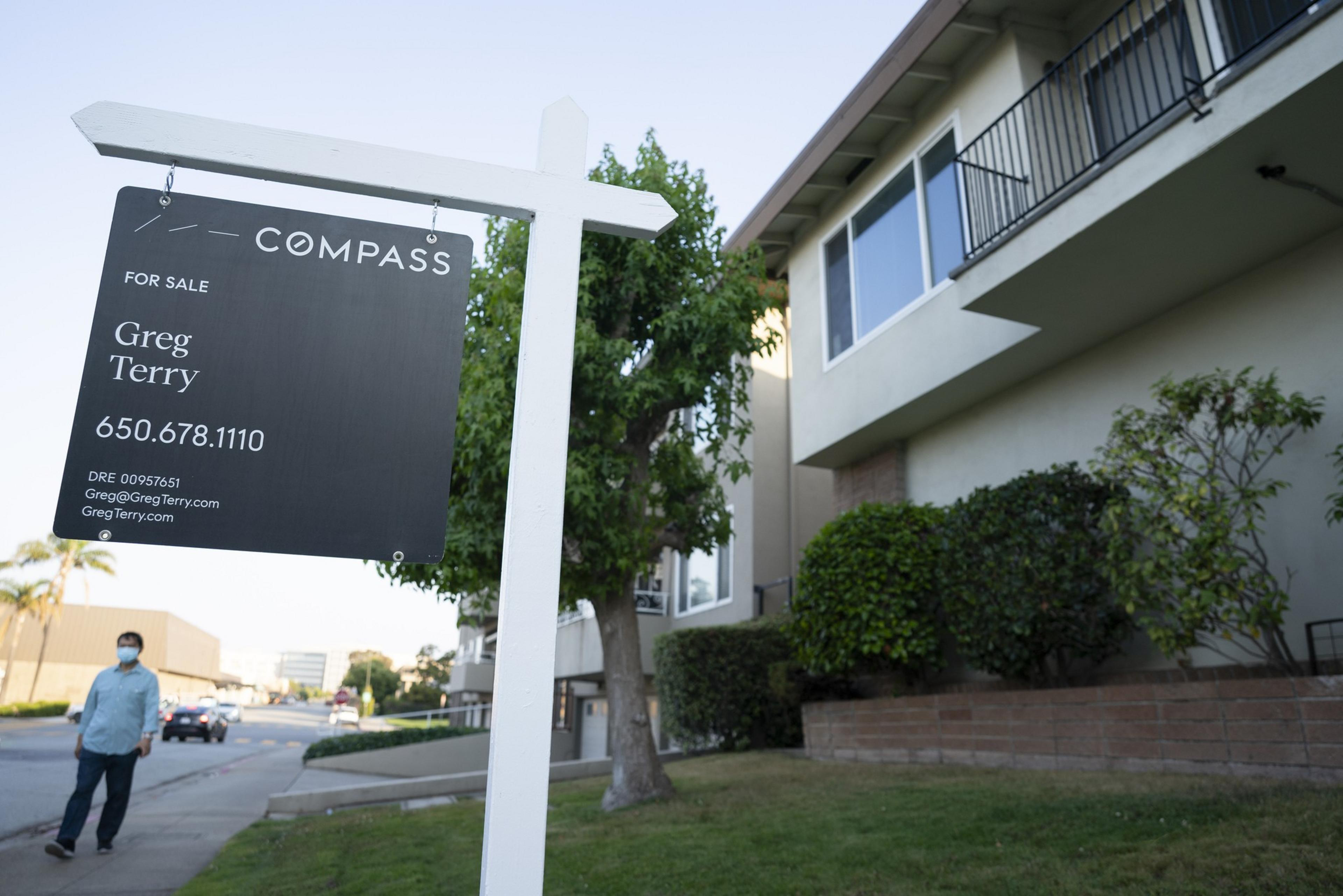 A &quot;For Sale&quot; sign is placed in front of a multi-story building with lush greenery. A person wearing a mask walks nearby on the sidewalk.