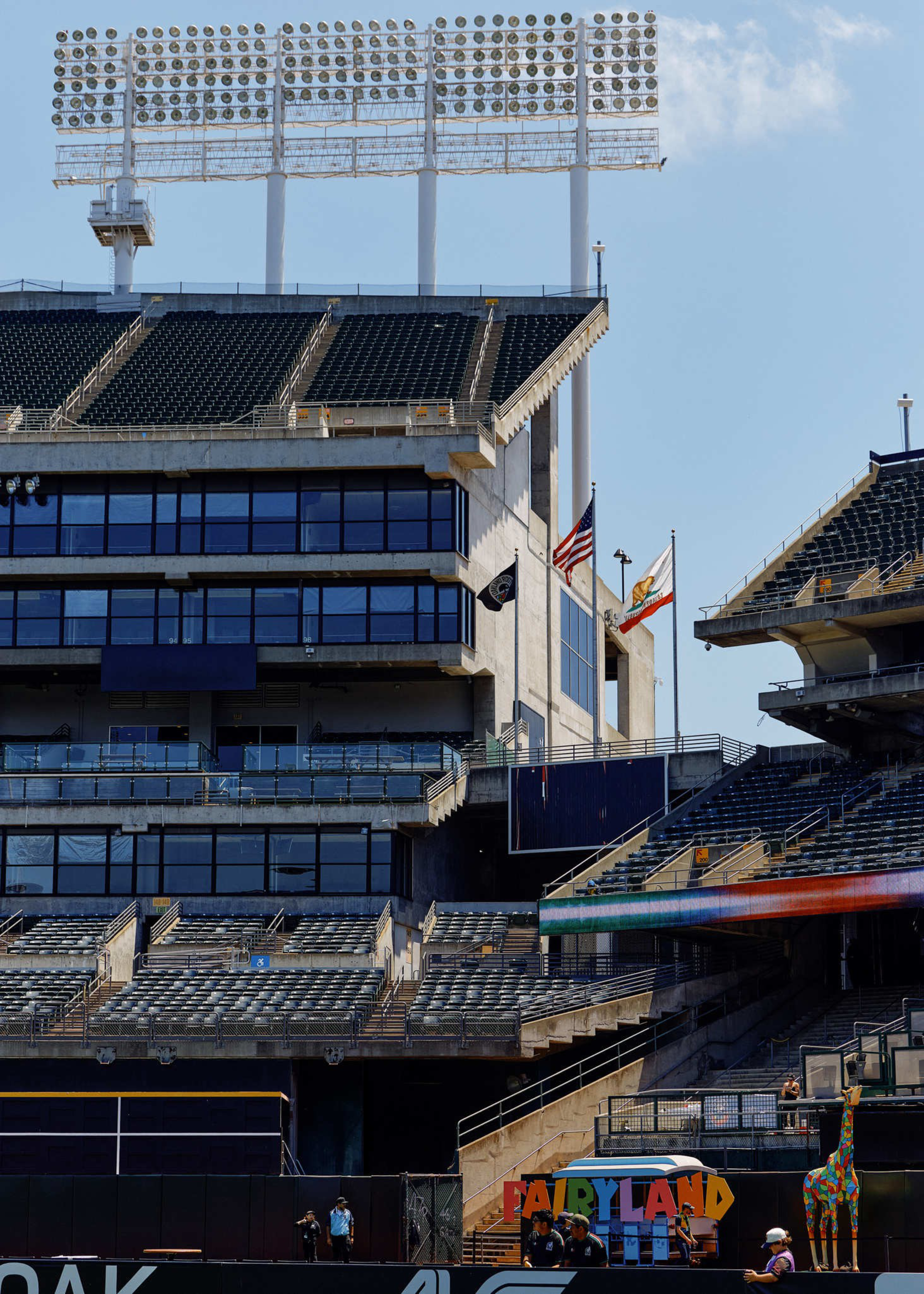 A mostly empty sports stadium with tiered seating, large floodlights above, three flags flying, and a colorful “Fairyland” sign with a giraffe figure near a few people.