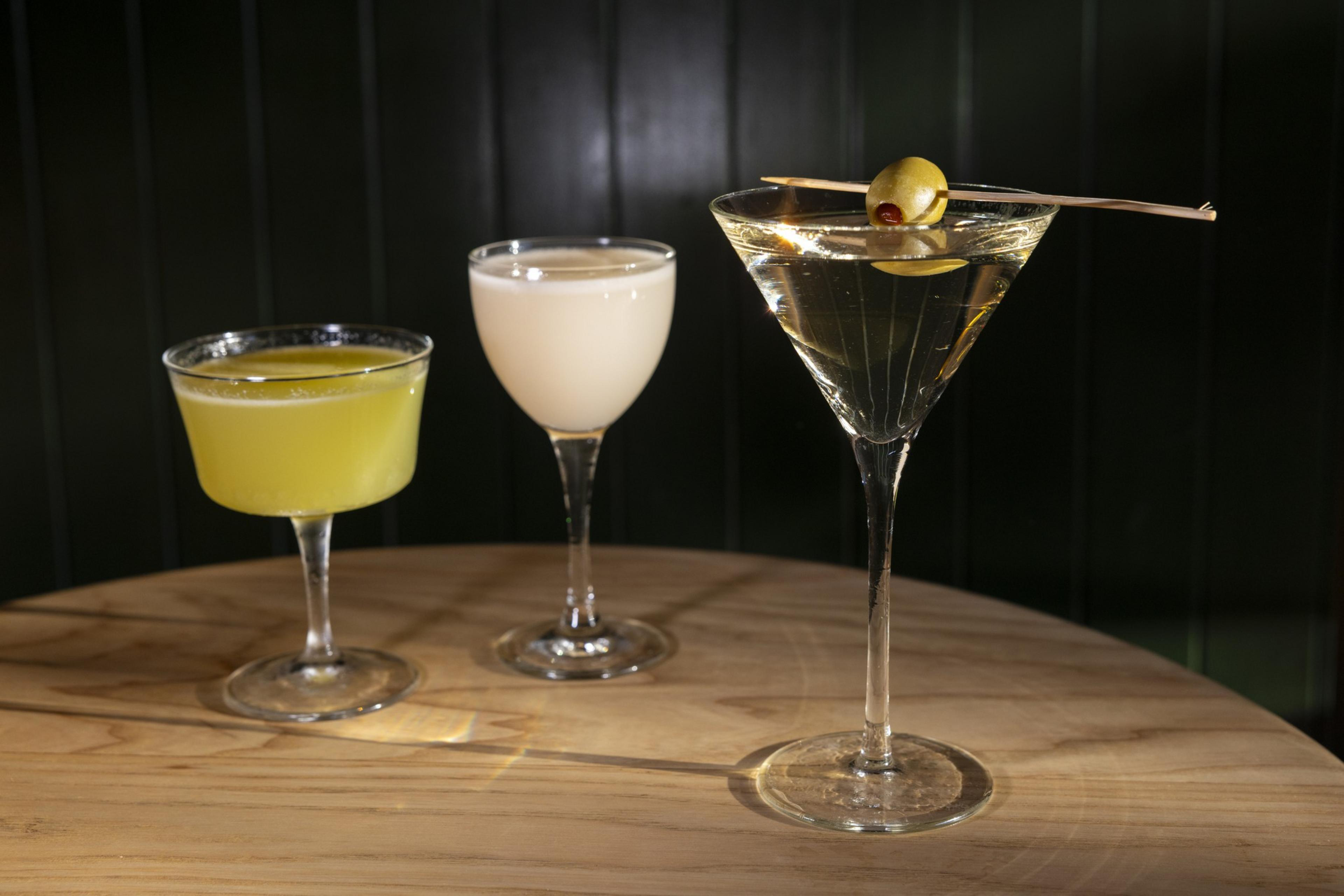 The image shows three cocktails on a round wooden table: a yellow drink in a coupe glass, a creamy drink in a stemmed glass, and a martini with an olive garnish.