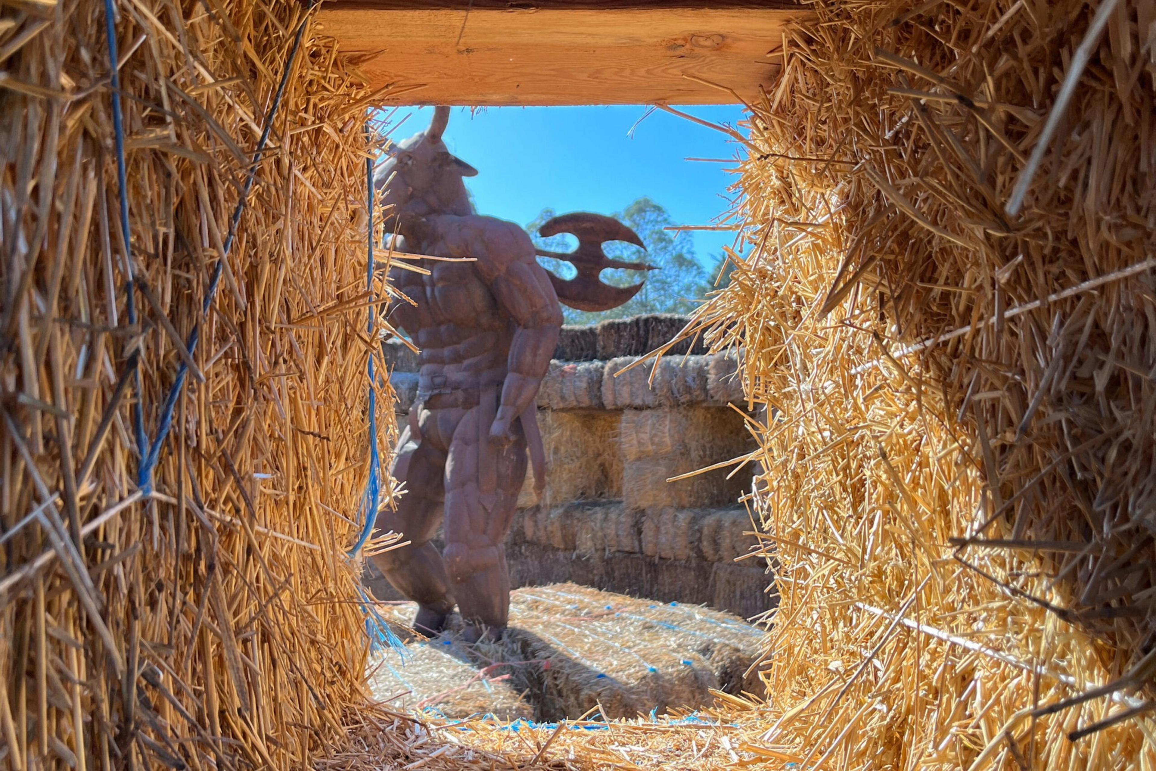 A muscular minotaur statue holding a large double-headed axe stands among rectangular hay bales under a clear blue sky.