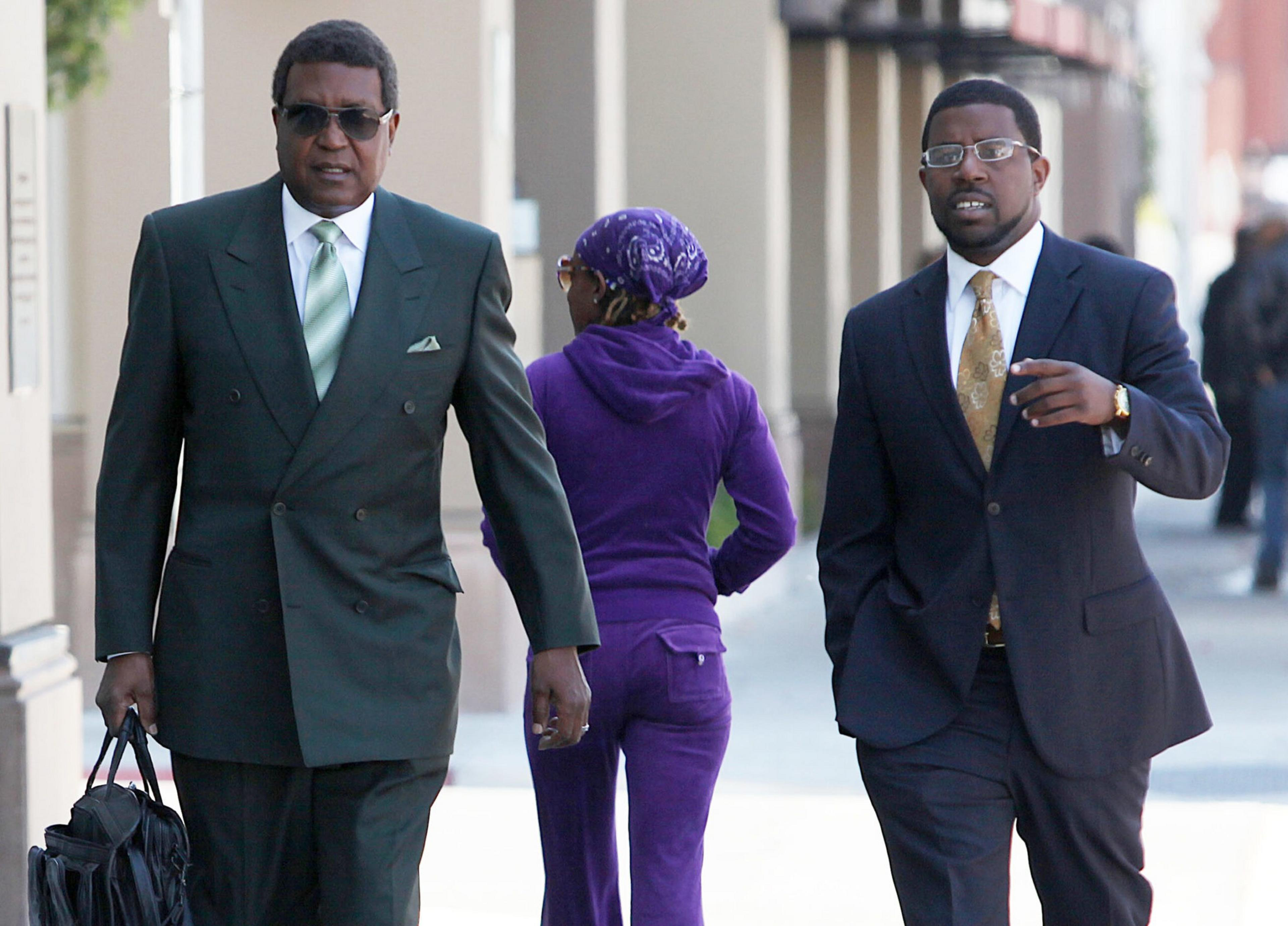 Two men in suits walk on a sidewalk, one carrying a black bag, while a woman in a purple tracksuit and headscarf walks away between them.