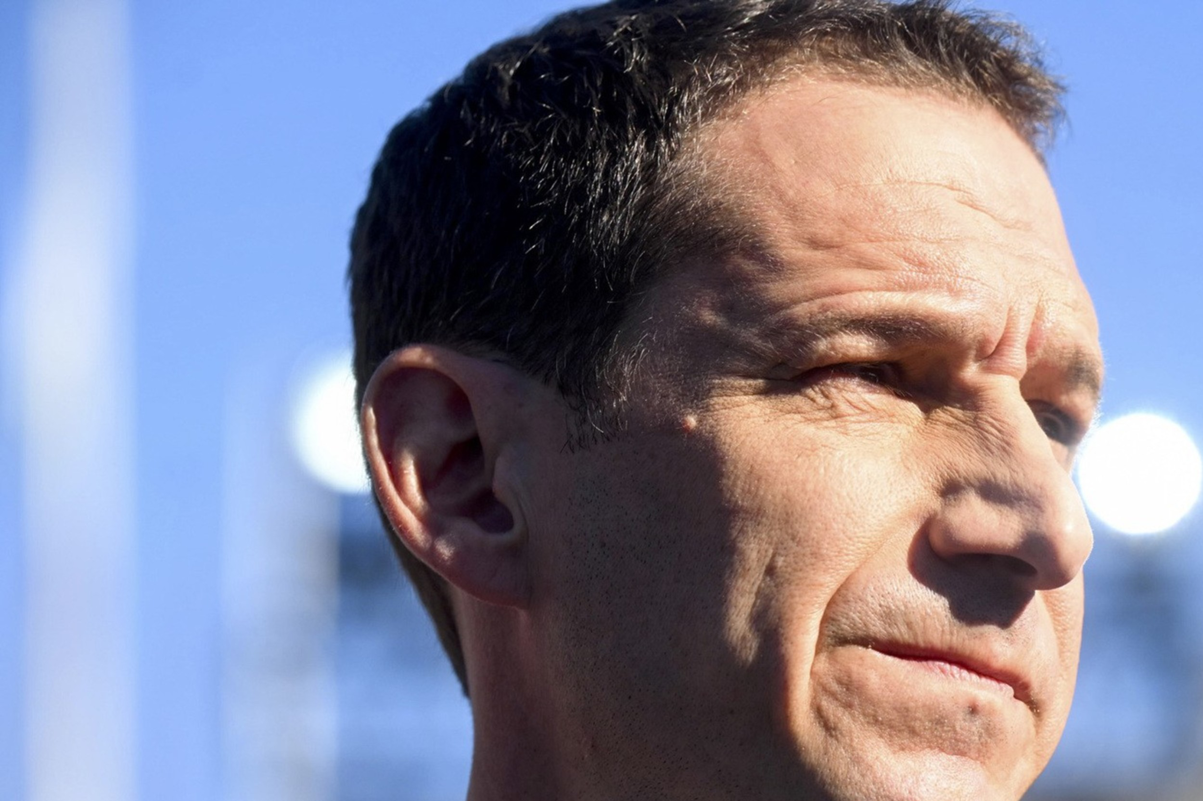 A man in a suit and tie stands outdoors with a focused expression against a clear blue sky, partially illuminated by bright sunlight.