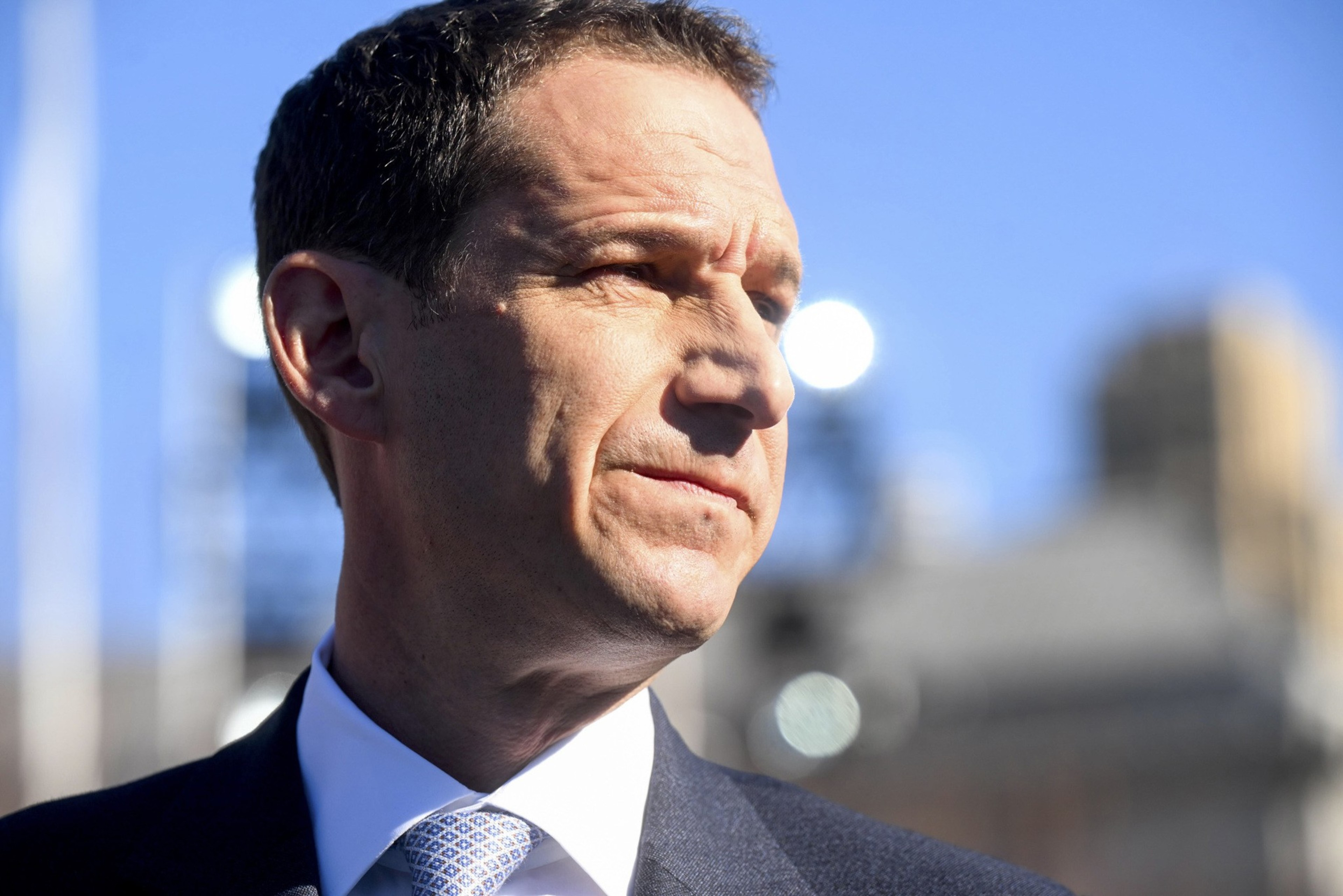 A man in a suit and tie stands outdoors with a focused expression against a clear blue sky, partially illuminated by bright sunlight.
