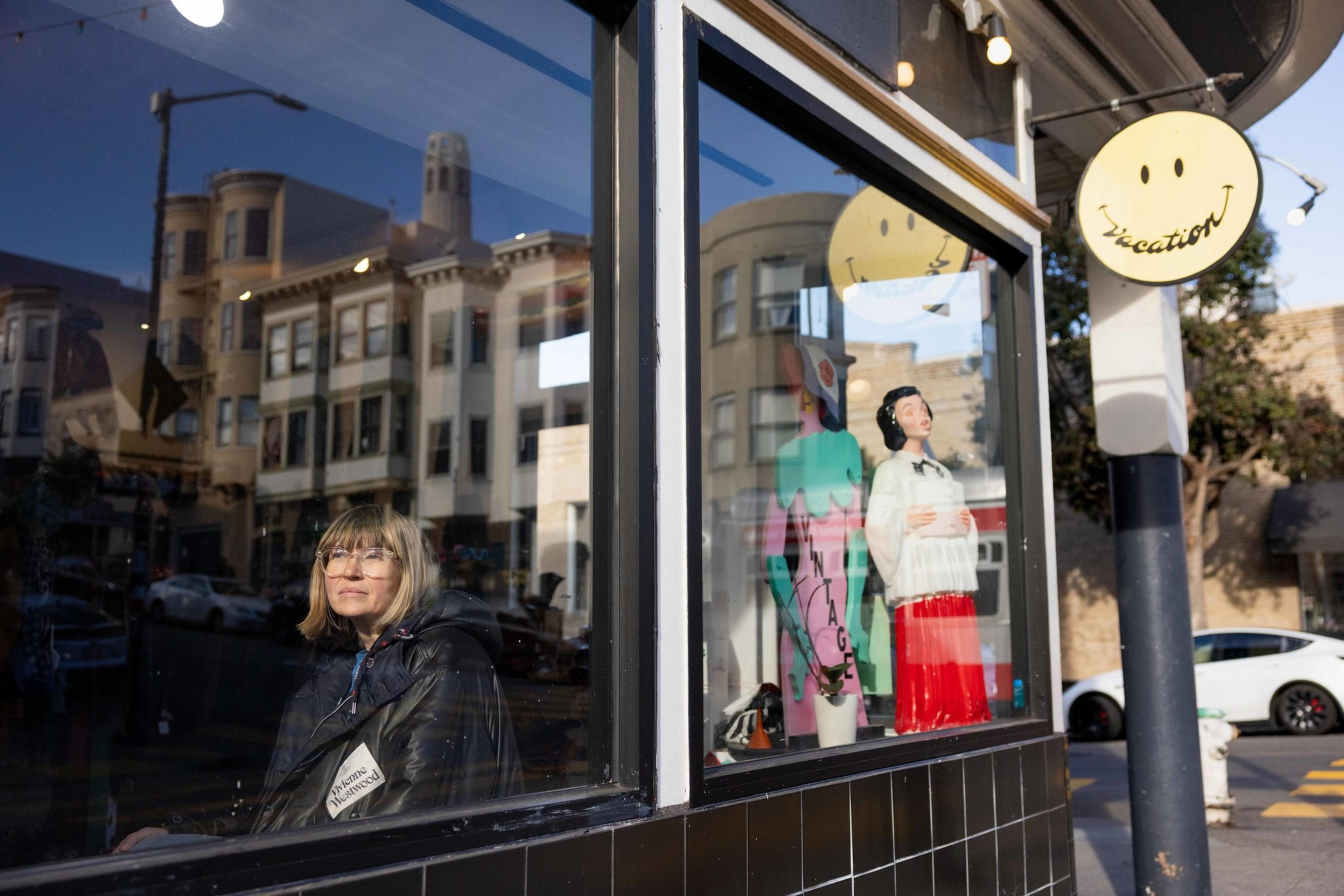 A woman is sitting inside, looking out a window of a shop with vintage decor. A reflection shows buildings and a smiling face sign reads &quot;Vacation.&quot;