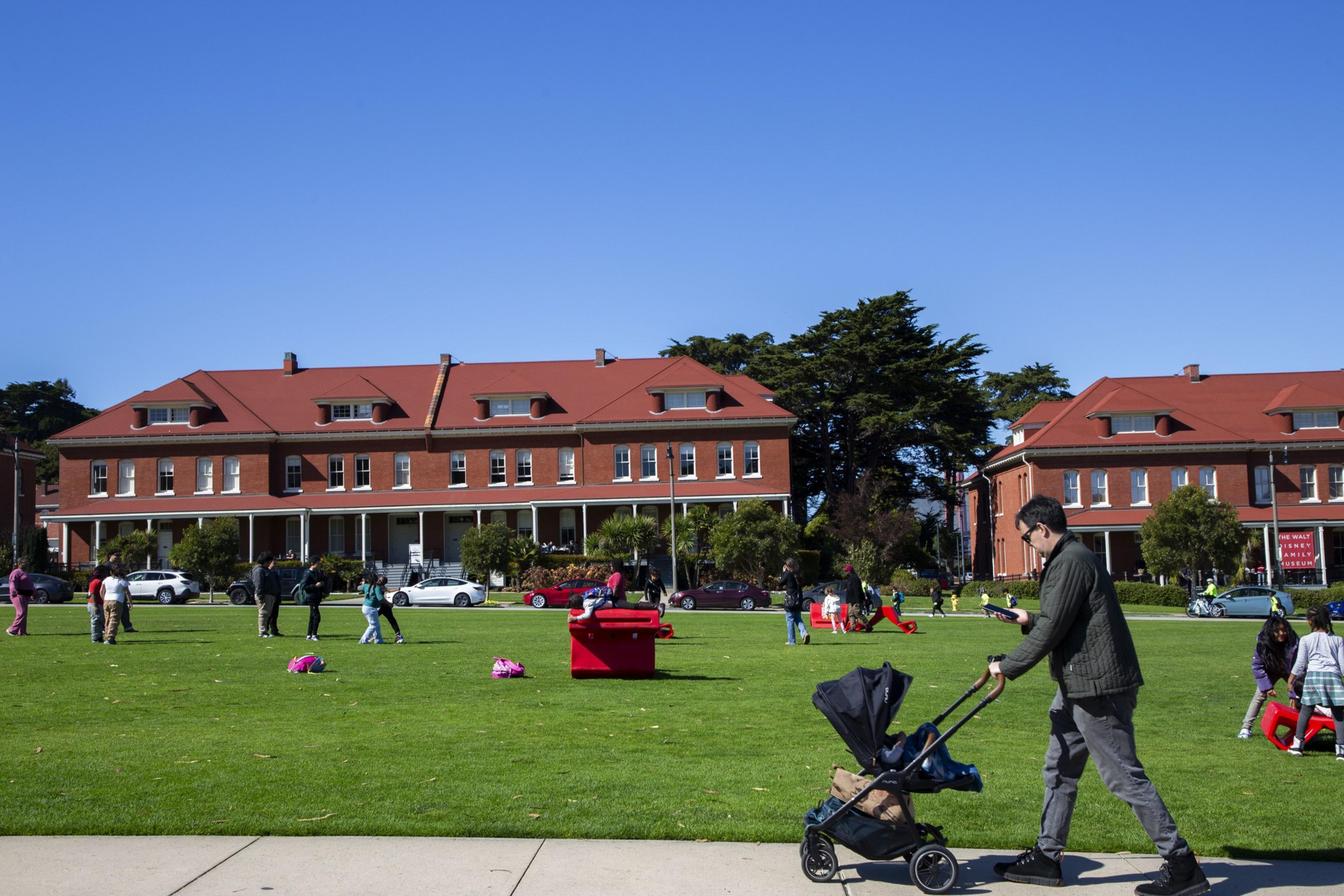 A man pushes a stroller across a grassy field, where people are gathered and children play. Red brick buildings with red roofs line the background under a clear blue sky.