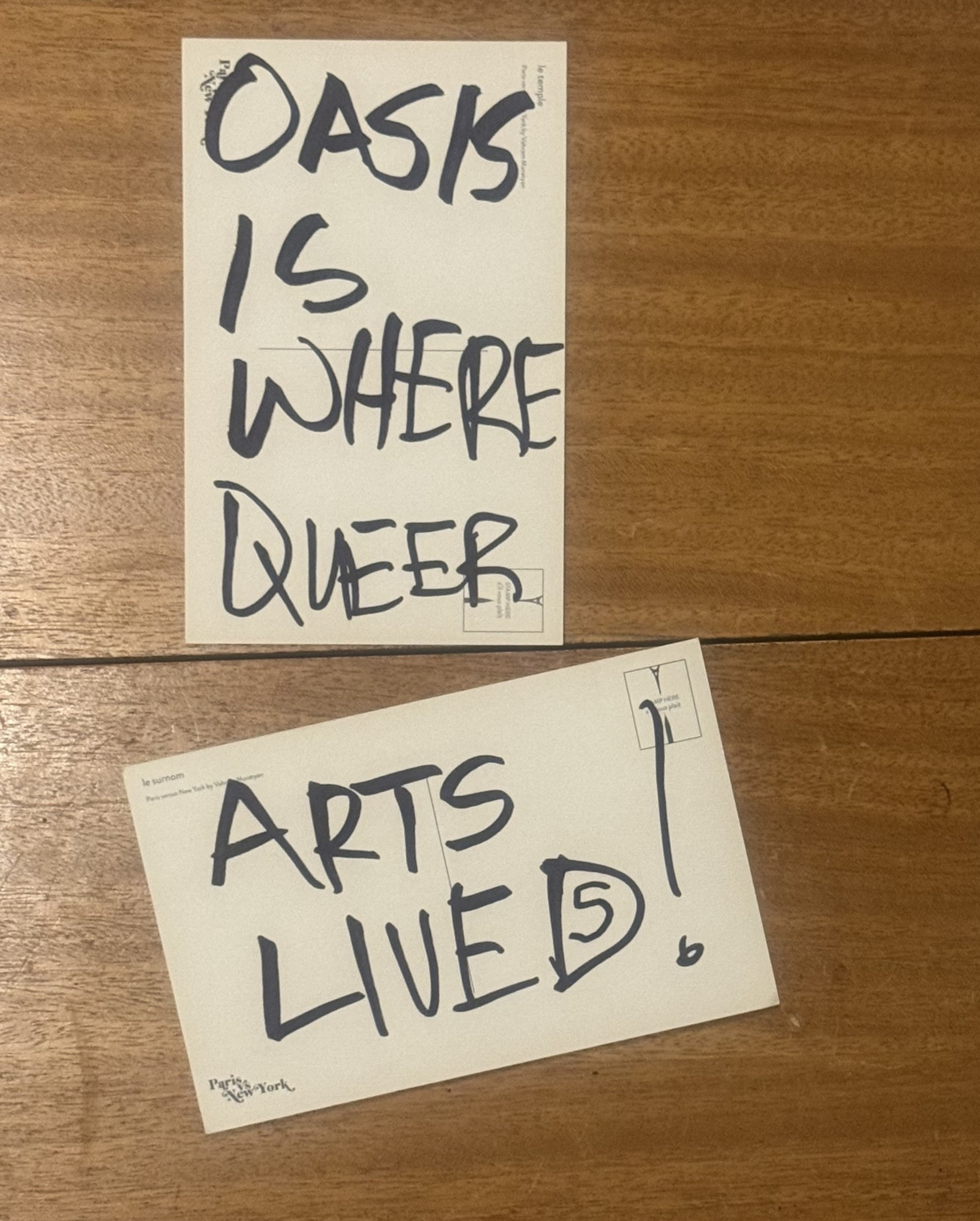 Oasis is where queer arts live, written in bold black letters on two postcards placed on a wooden surface.
