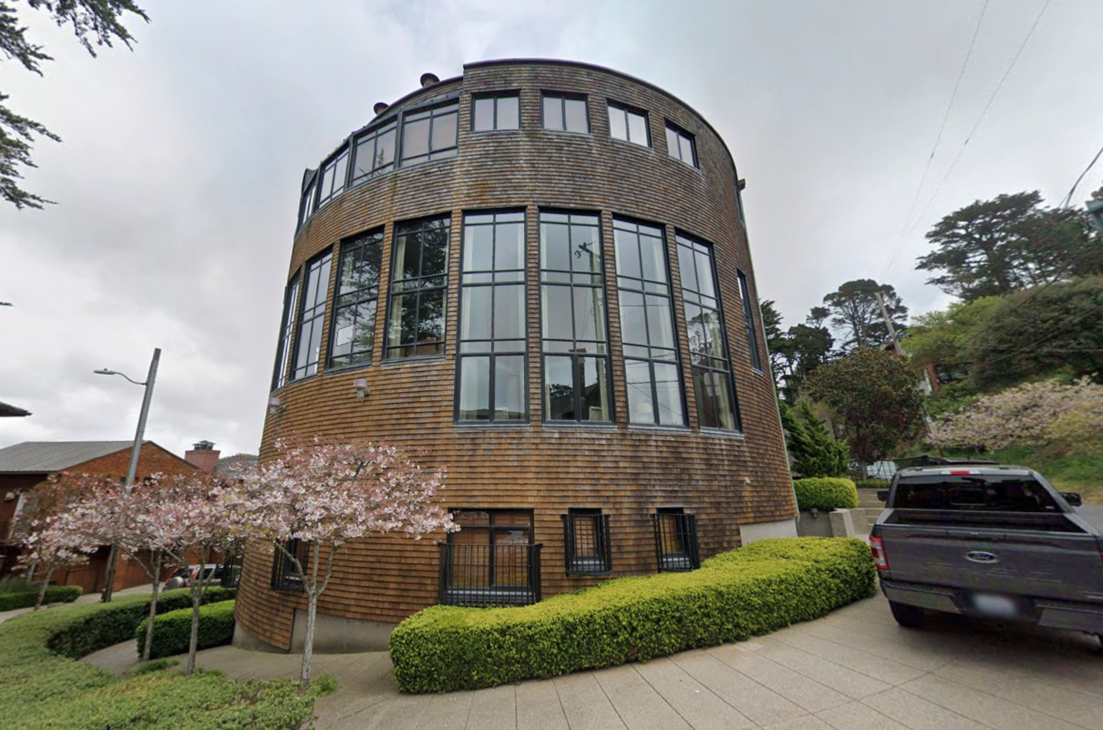 A three-story cylindrical wooden building with large windows stands on a corner, surrounded by trimmed bushes and a sidewalk with a parked gray pickup truck.