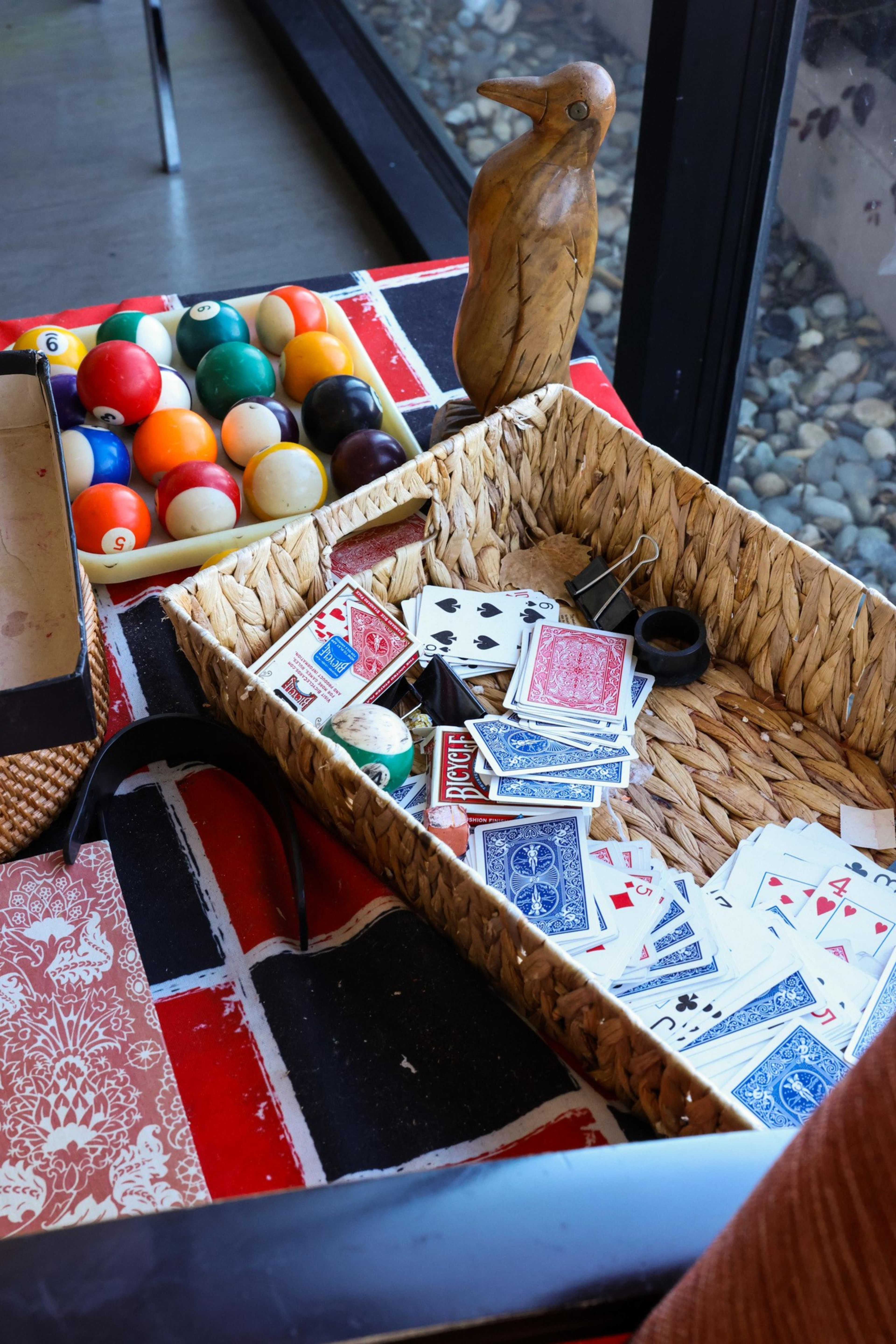 A basket on a patterned table holds scattered playing cards and a dark ring. Nearby are colorful billiard balls and a wooden bird sculpture.