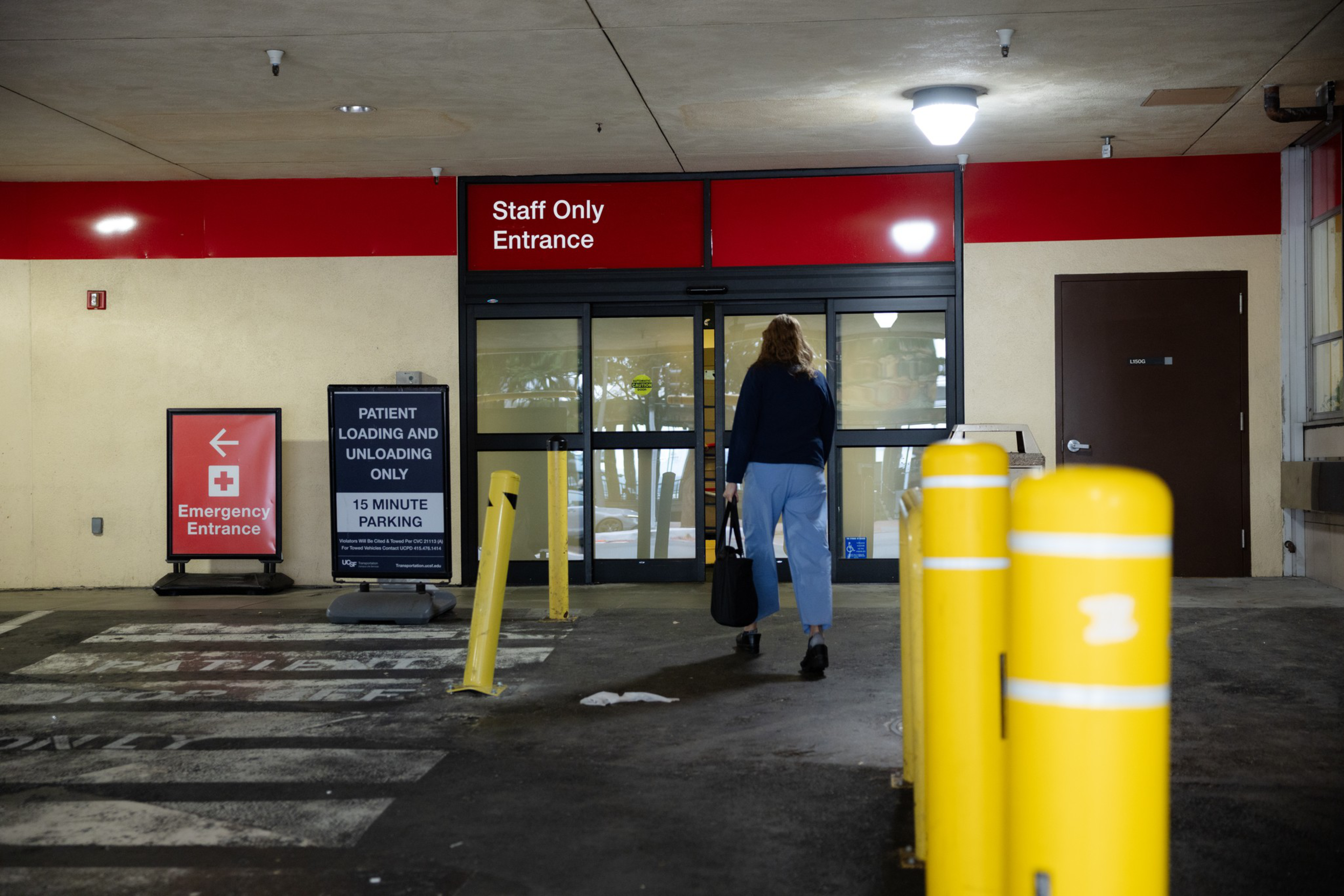 A person wearing blue pants walks toward a “Staff Only Entrance” door, with yellow bollards nearby and signs indicating emergency entrance and patient loading.