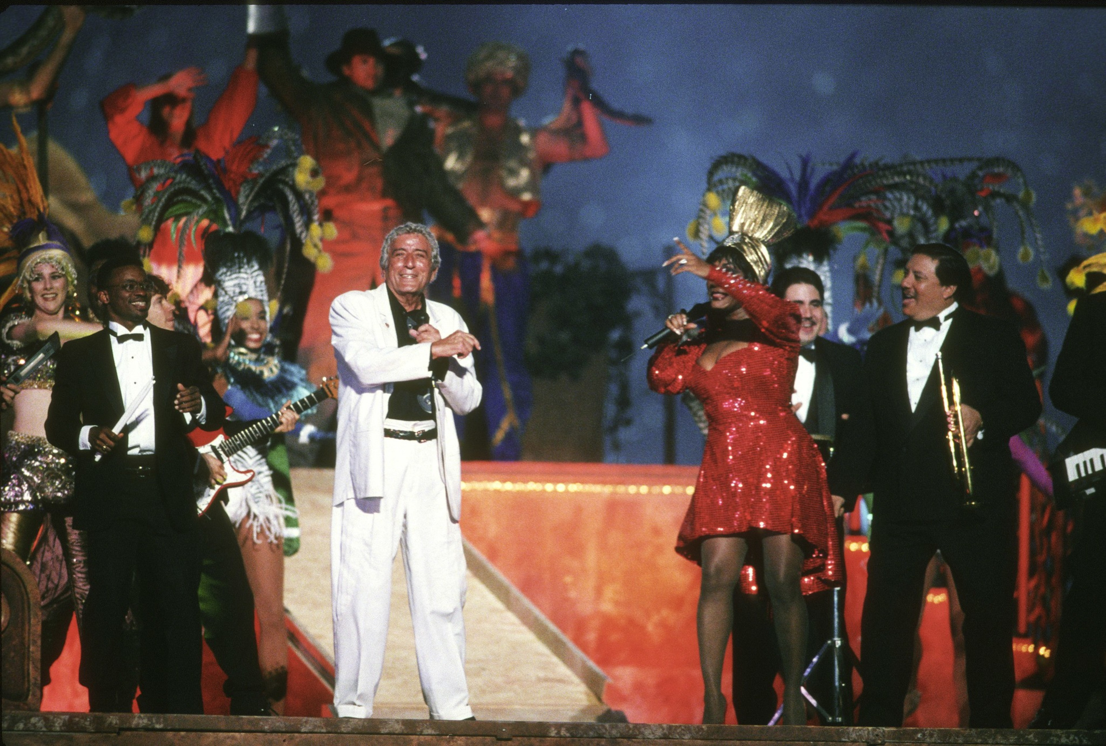 Performers in tuxedos, a white suit, and colorful costumes sing and play instruments on a stage decorated with feathers and vibrant lights.