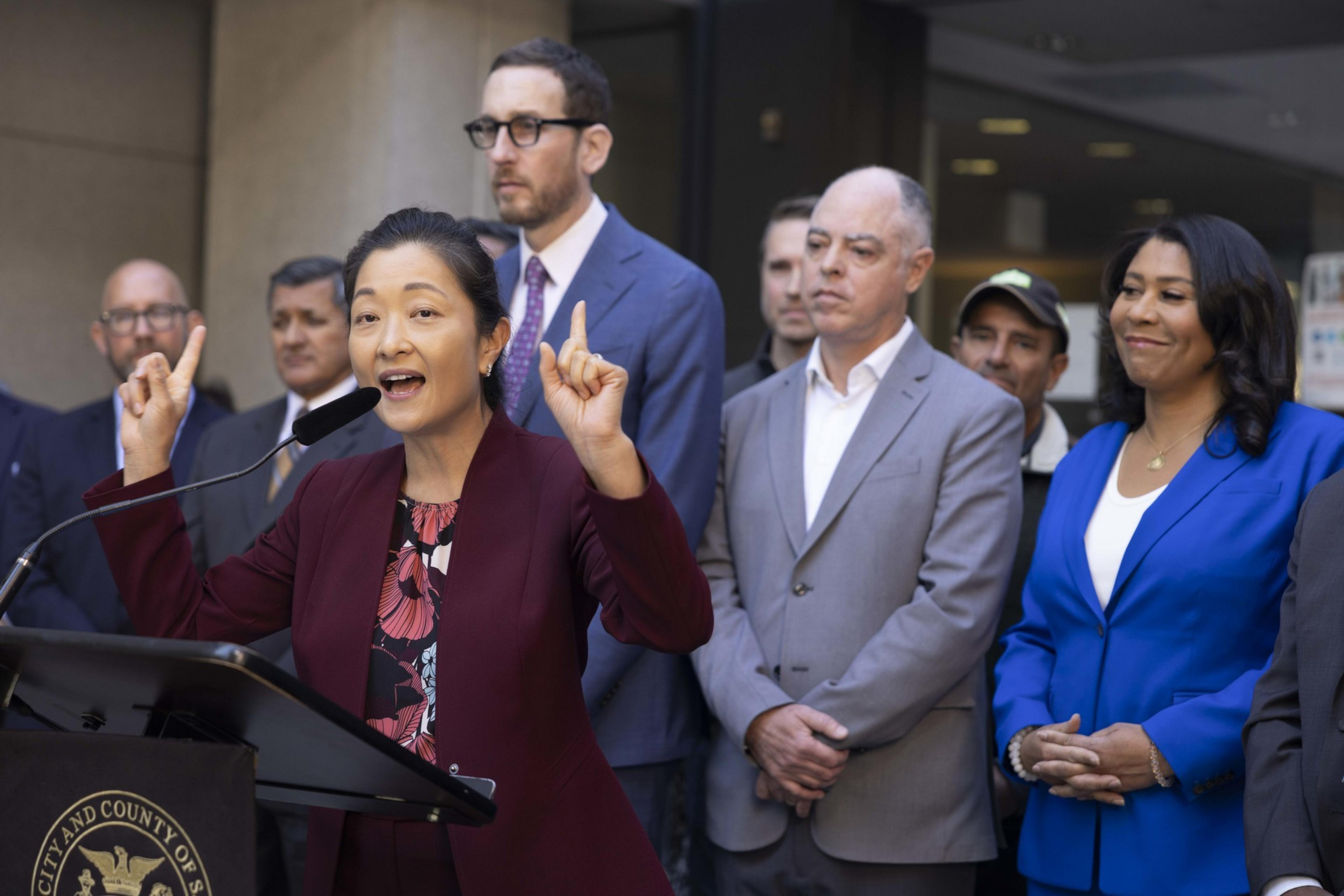 A woman in a maroon suit speaks passionately at a podium, surrounded by several attentive people in suits and a woman in a blue blazer.