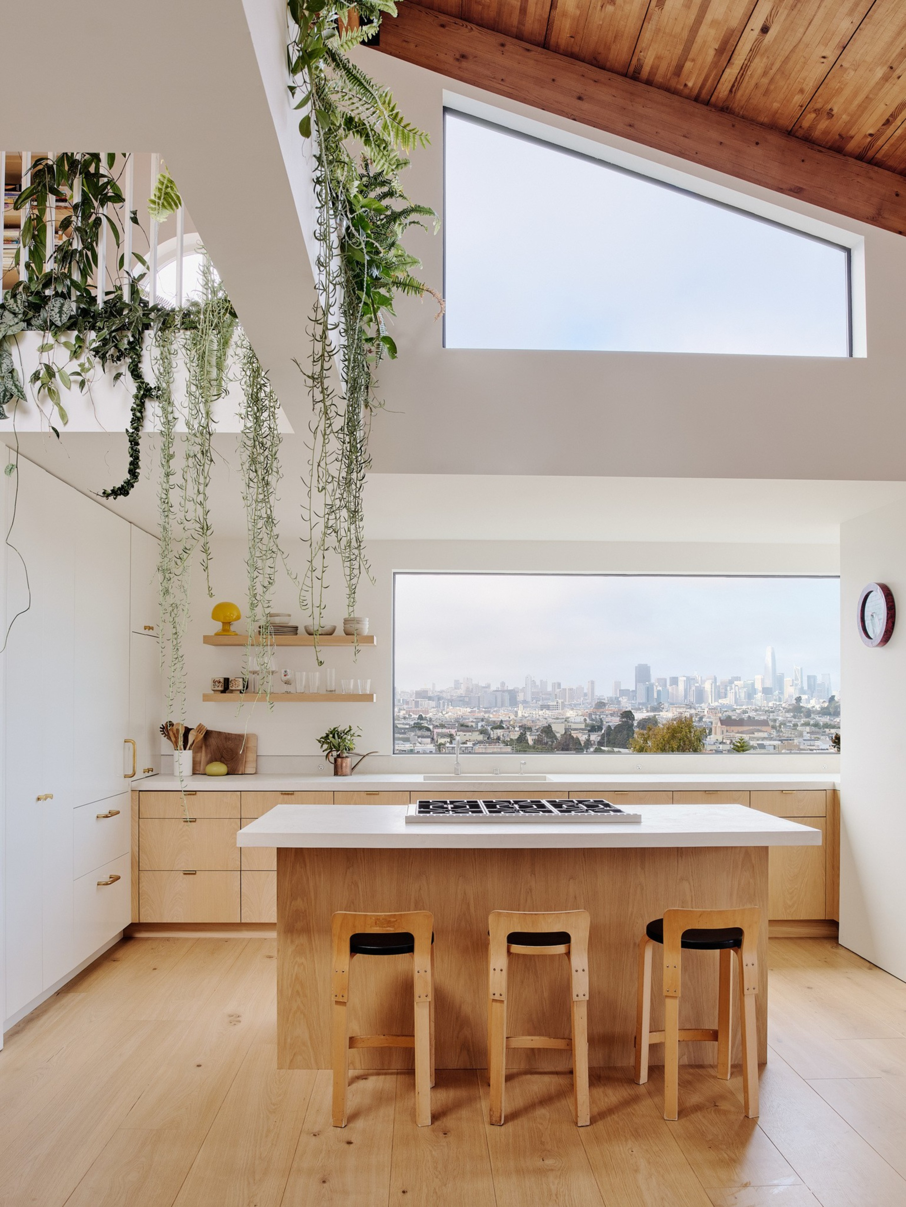 The image shows a modern kitchen with a wooden island, three stools, hanging plants, a large window with a city view, and a skylight above.