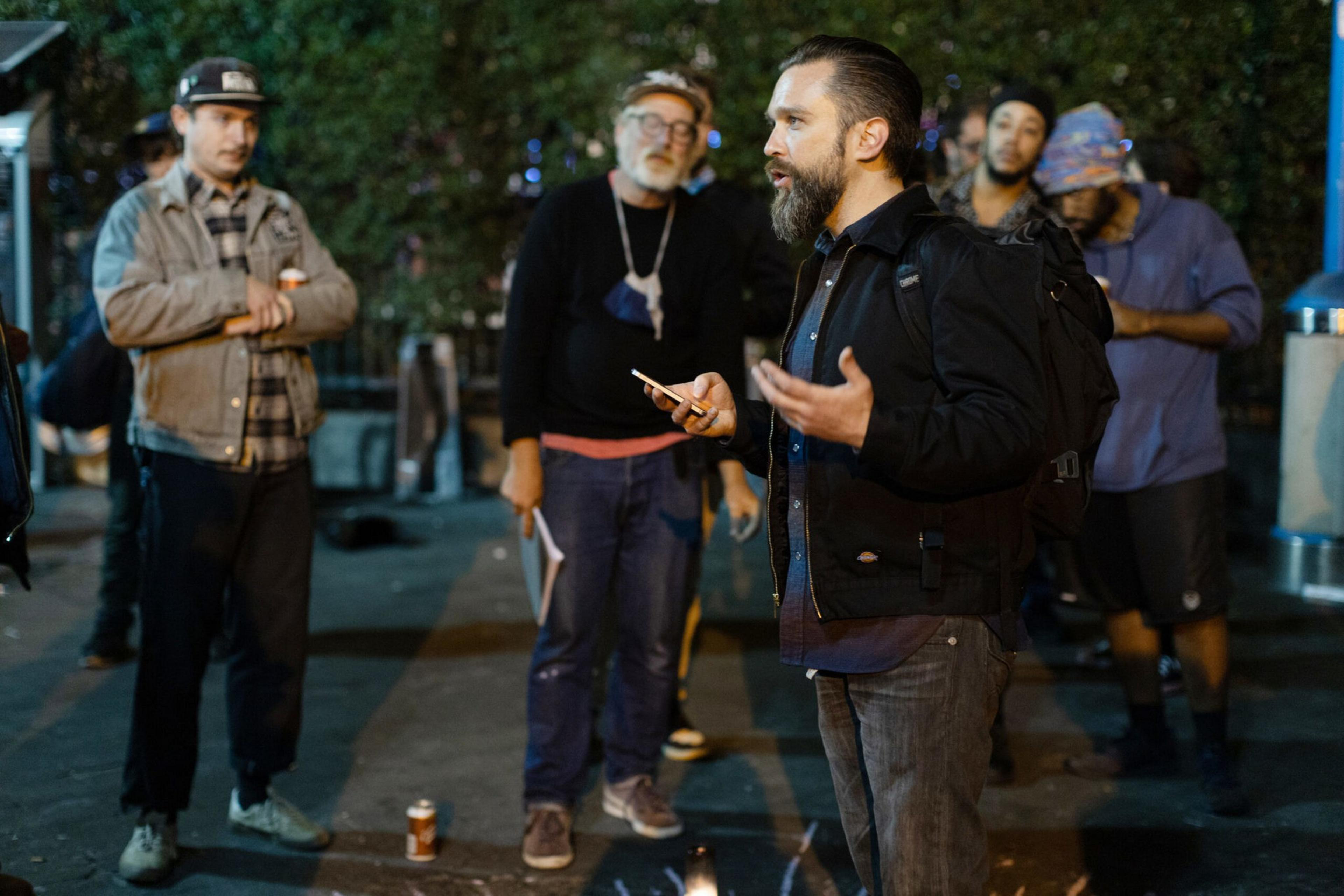 A man with a beard speaks animatedly to a small group of casually dressed people standing outdoors at night.