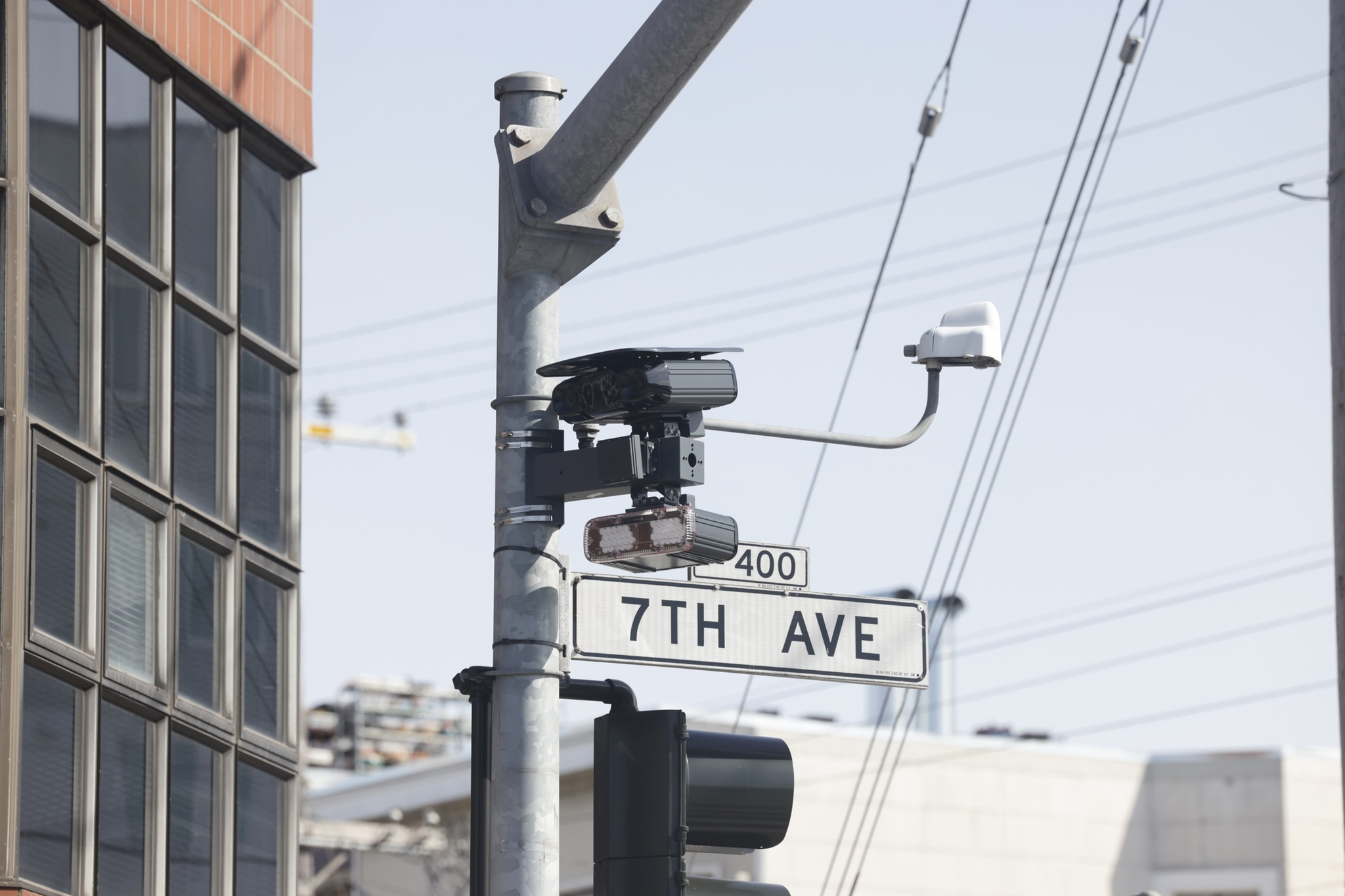A street sign labeled "7TH AVE" is mounted on a pole with traffic cameras and a traffic light nearby, against a background of buildings and power lines.
