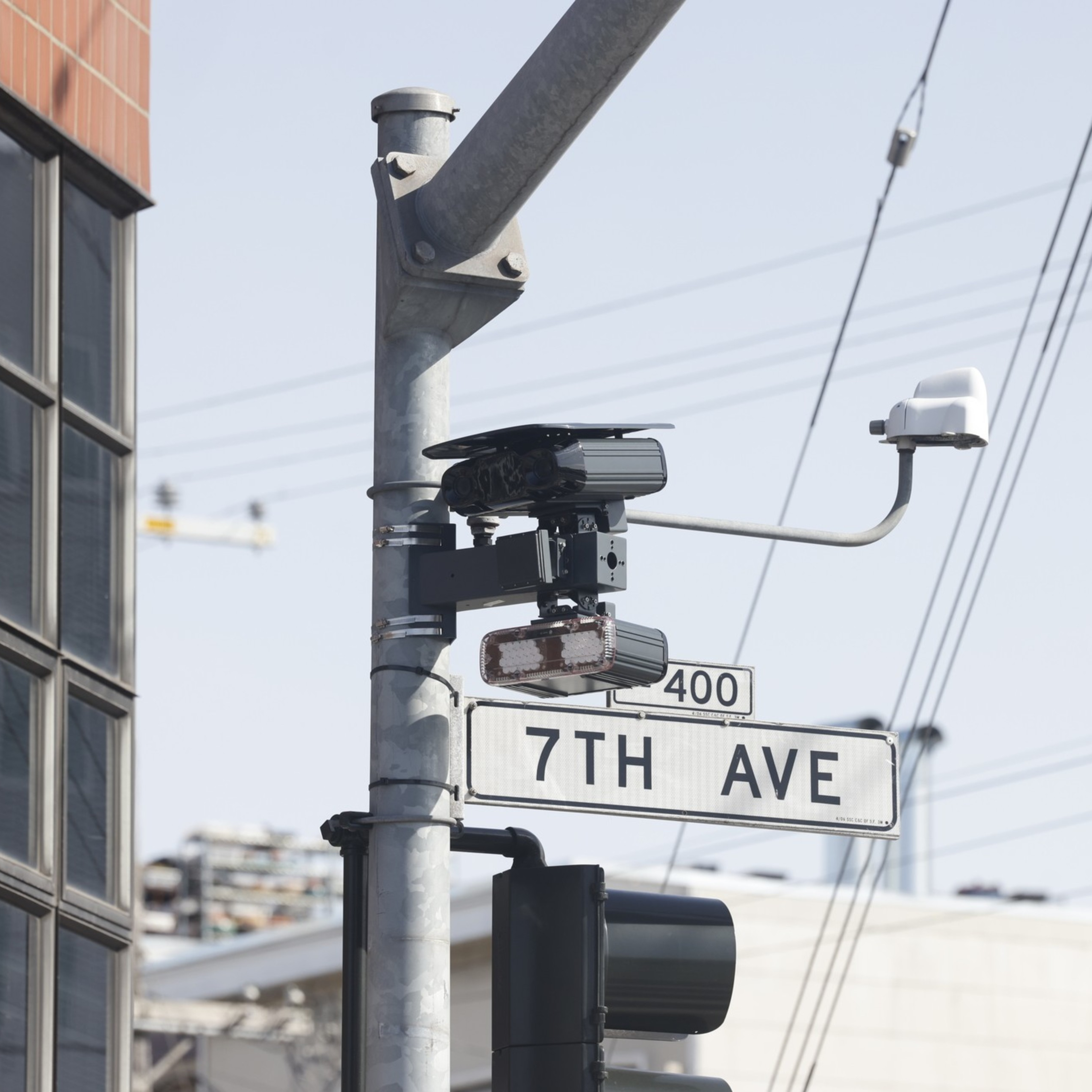 A street sign labeled "7TH AVE" is mounted on a pole with traffic cameras and a traffic light nearby, against a background of buildings and power lines.