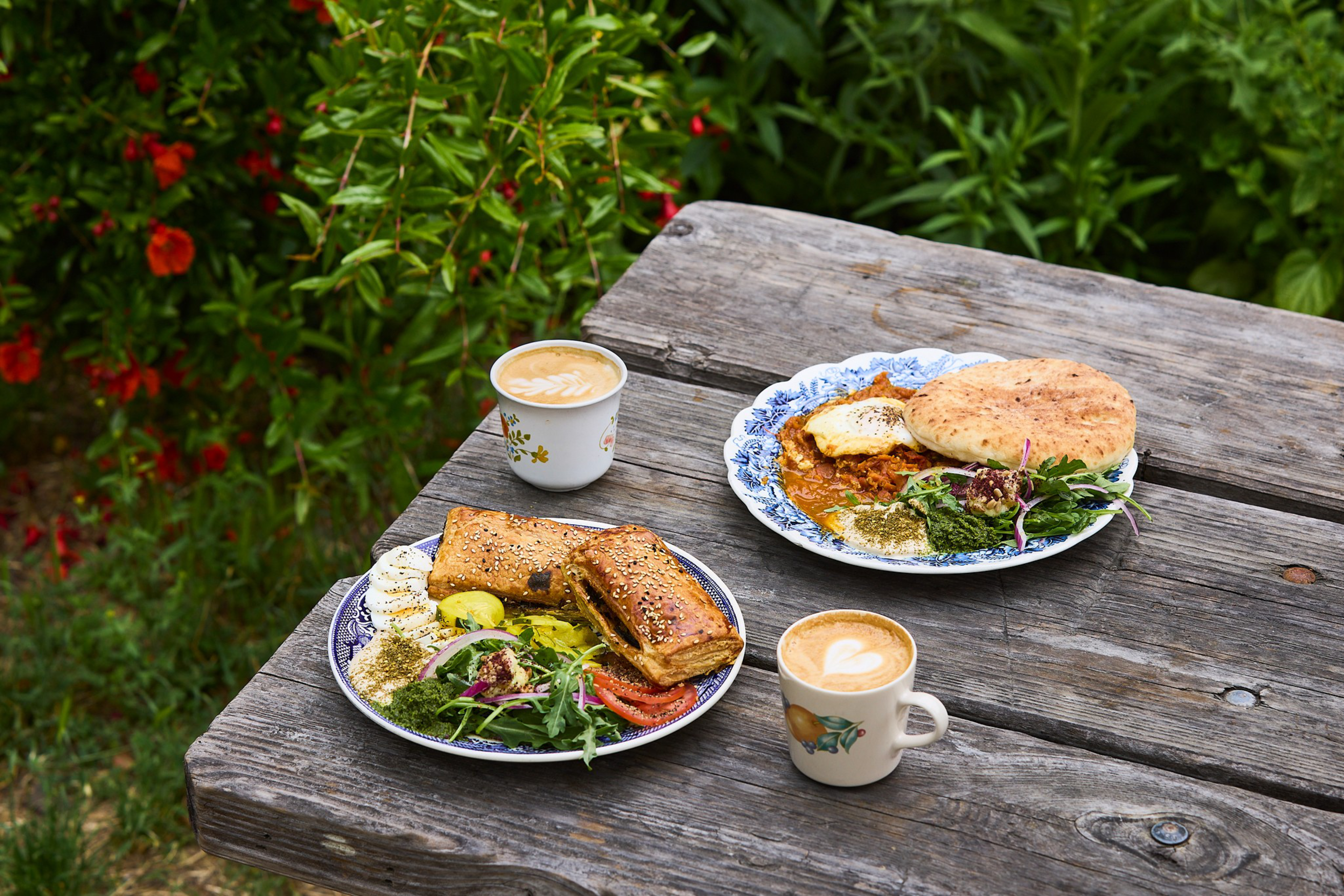 Two plates with savory pastries, salads, and dips sit on a weathered wooden table outdoors, accompanied by two cups of coffee with latte art.
