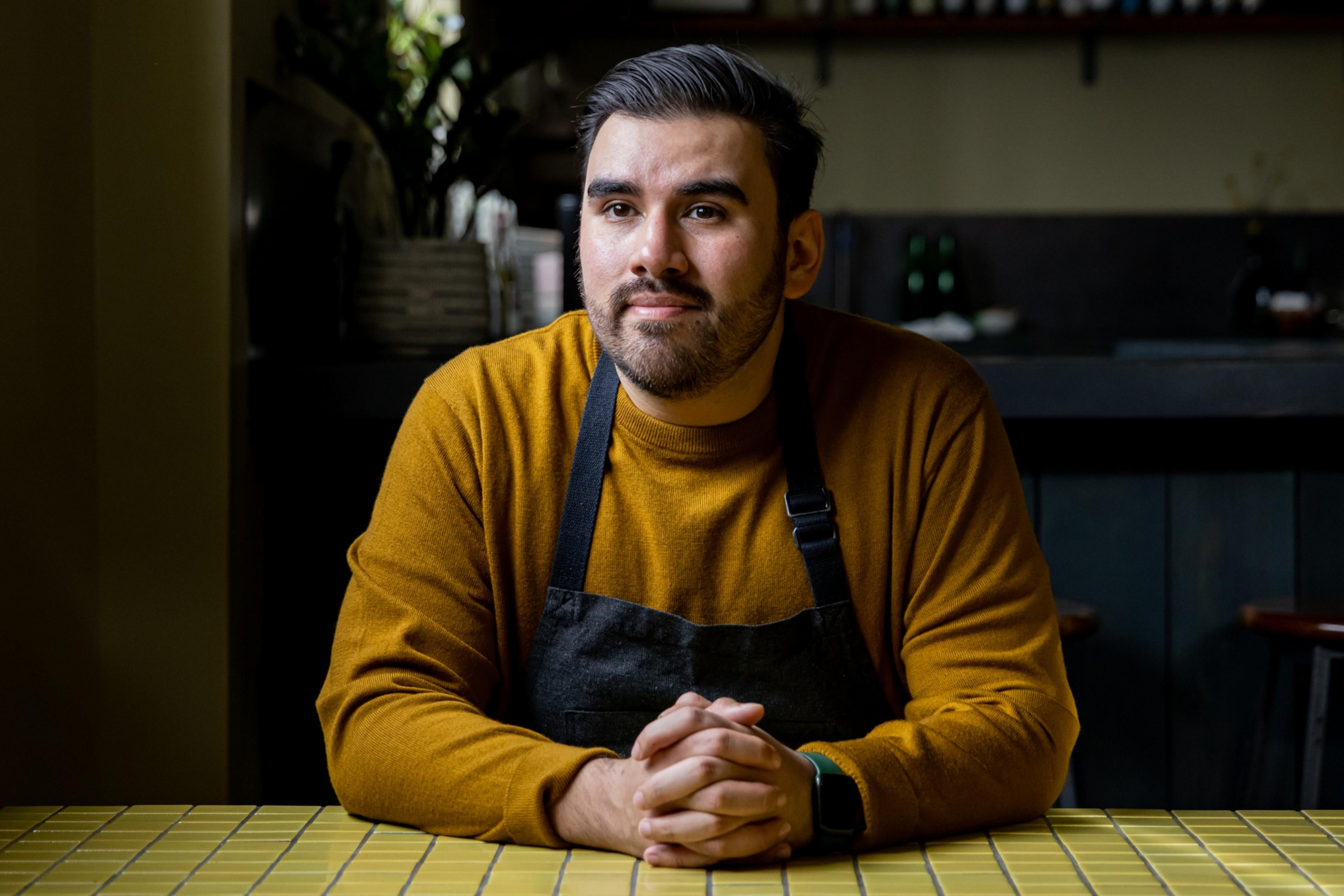 A man with a beard is sitting at a tiled table, wearing a mustard sweater and a black apron. The background is a dimly lit interior with shelves and greenery.
