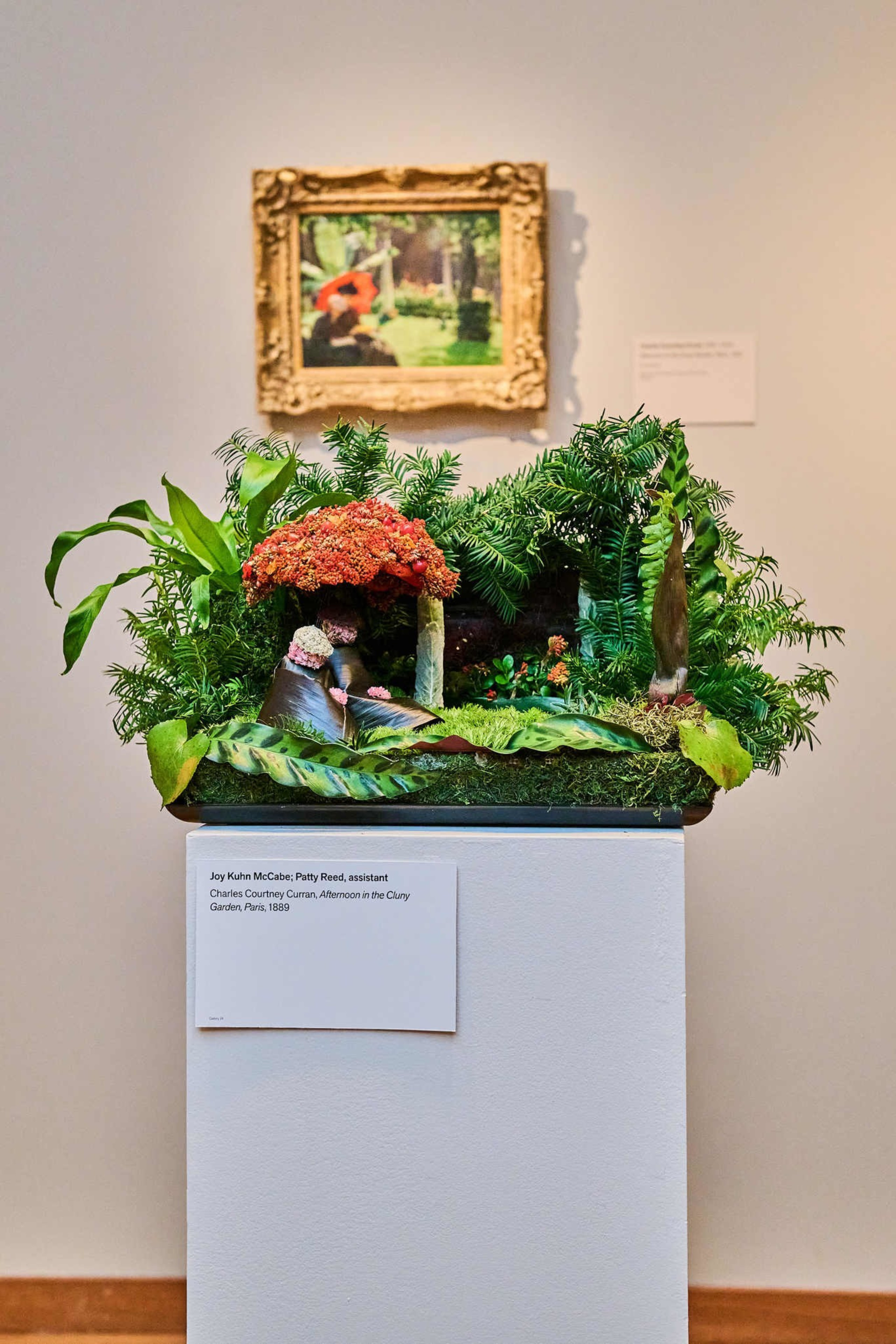 The image shows a vibrant floral arrangement mimicking a painting in a gold frame on the wall behind it. The floral display is lush and colorful with various plants.