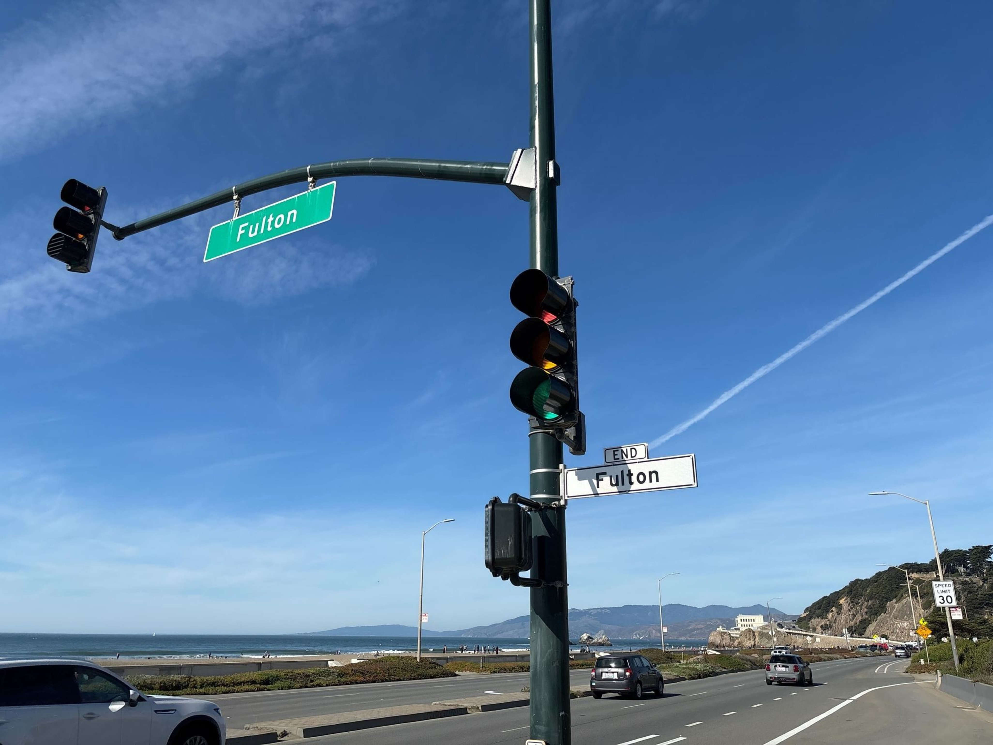 A traffic light at the end of Fulton street overlooks a coastal road with cars, a beach on the left, and hills in the distance under a clear blue sky.